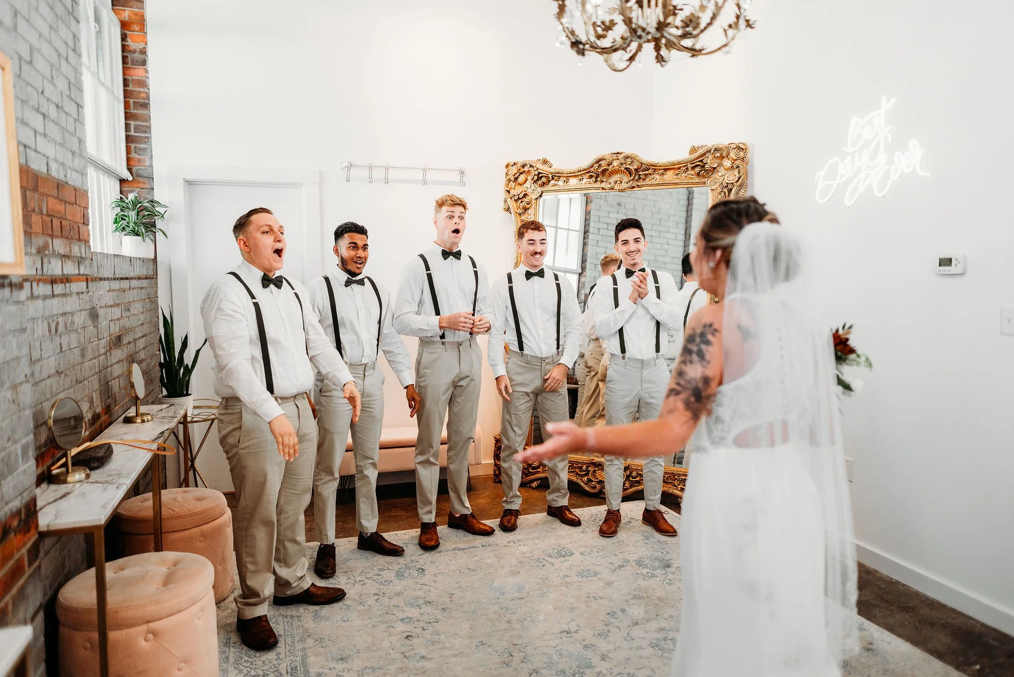 Bride in white dress and veil facing five groomsmen in white shirts, beige pants, suspenders, and bow ties, inside a decorated room with mirror and neon sign.