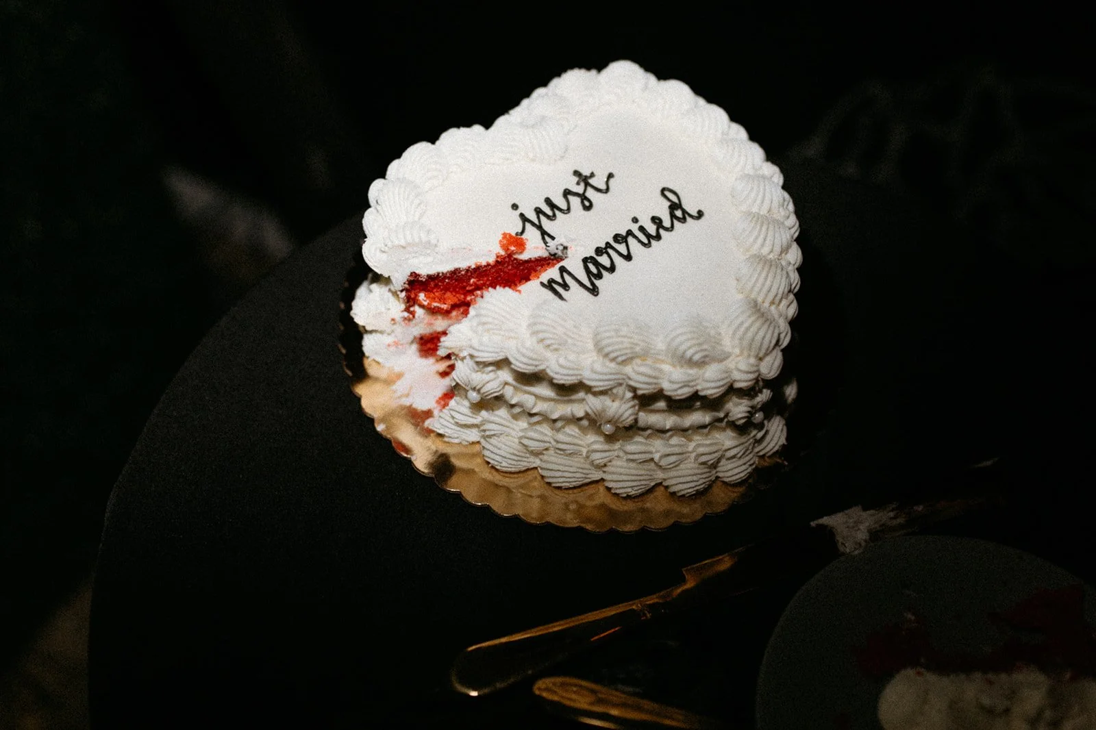 A heart-shaped slice of wedding cake with white icing and decorative piping, with a bite revealing red velvet layers, on a black surface with a gold-colored cake server.