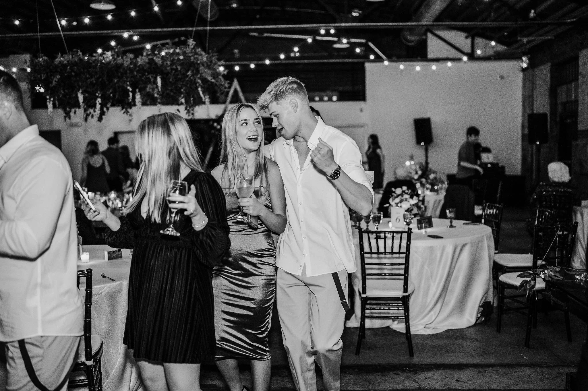 Three young adults, two women and one man, dancing and talking at a formal event with decorated tables and string lights overhead in a large indoor venue.
