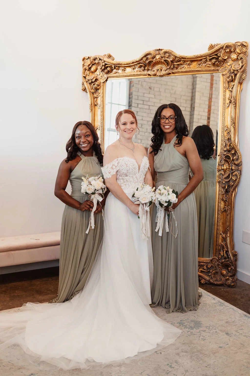 A bride and two bridesmaids standing in front of a large decorative mirror, smiling and holding bouquets, during a wedding.