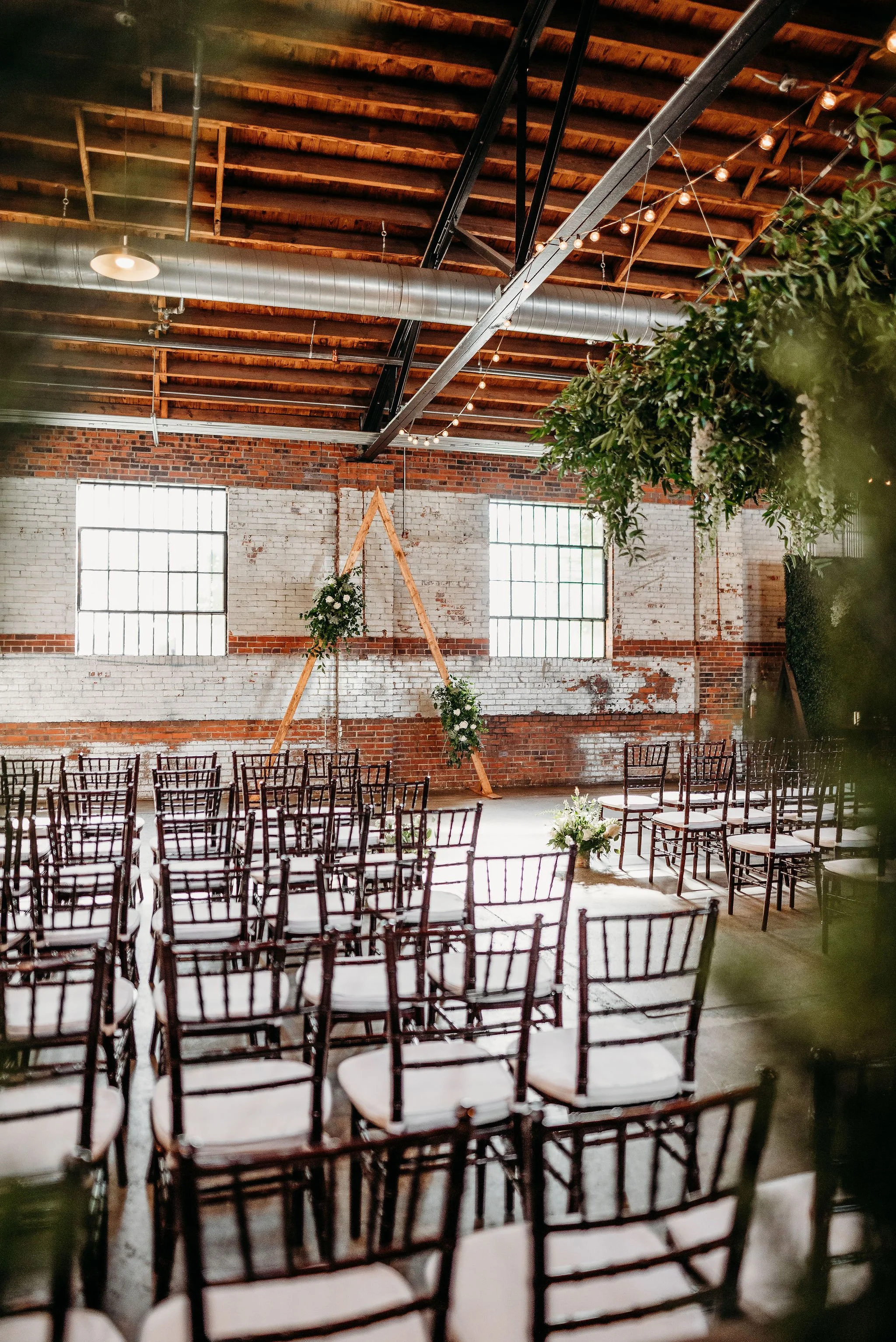 Empty chairs arranged in rows for a wedding ceremony inside a rustic industrial venue with exposed brick walls, large windows, and string lights. A triangular wooden arch decorated with greenery and flowers stands at the front.