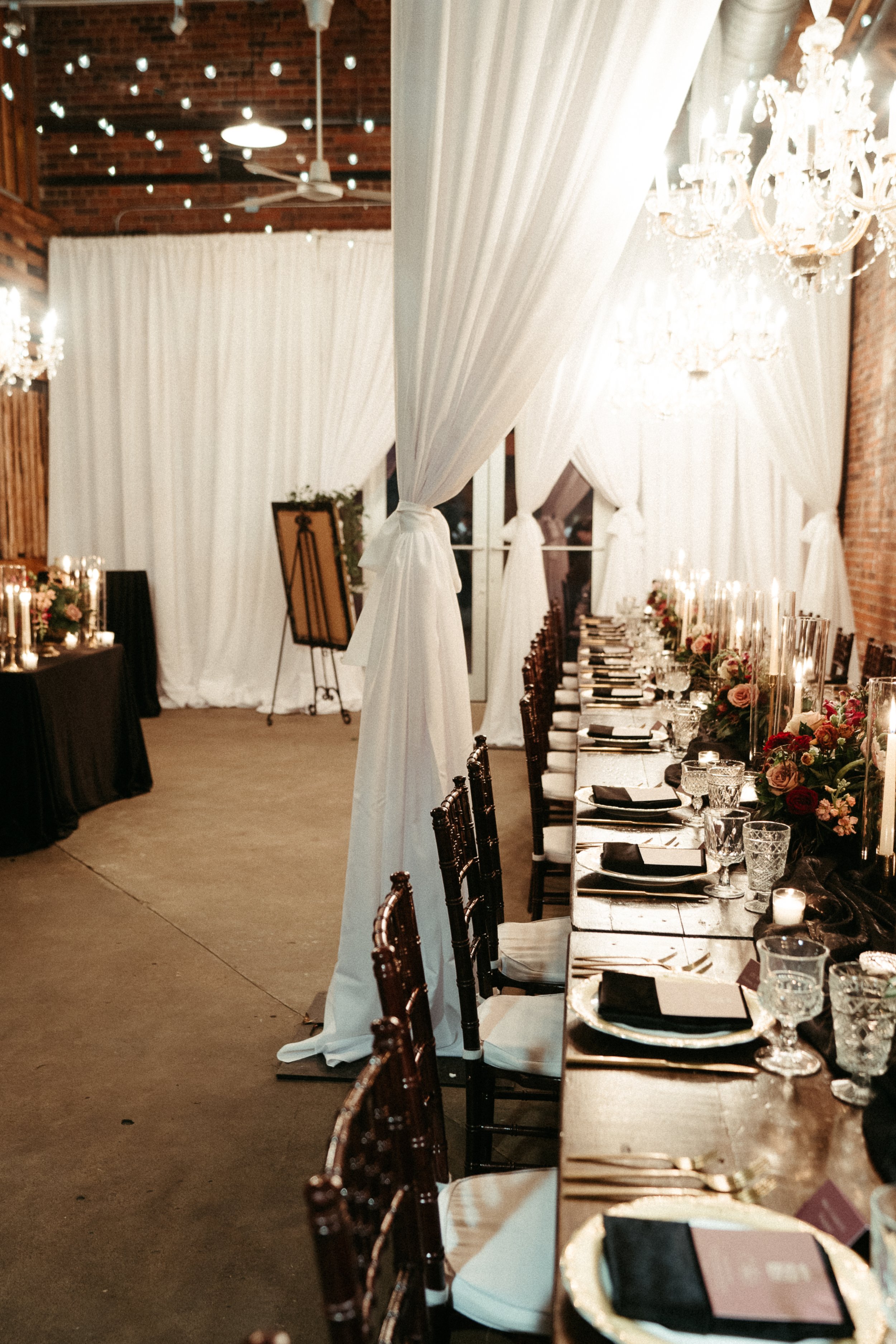 Elegant indoor wedding reception with a long table set with black napkins, glassware, floral centerpieces, and candles. White curtains and chandeliers decor the background, creating a romantic ambiance.