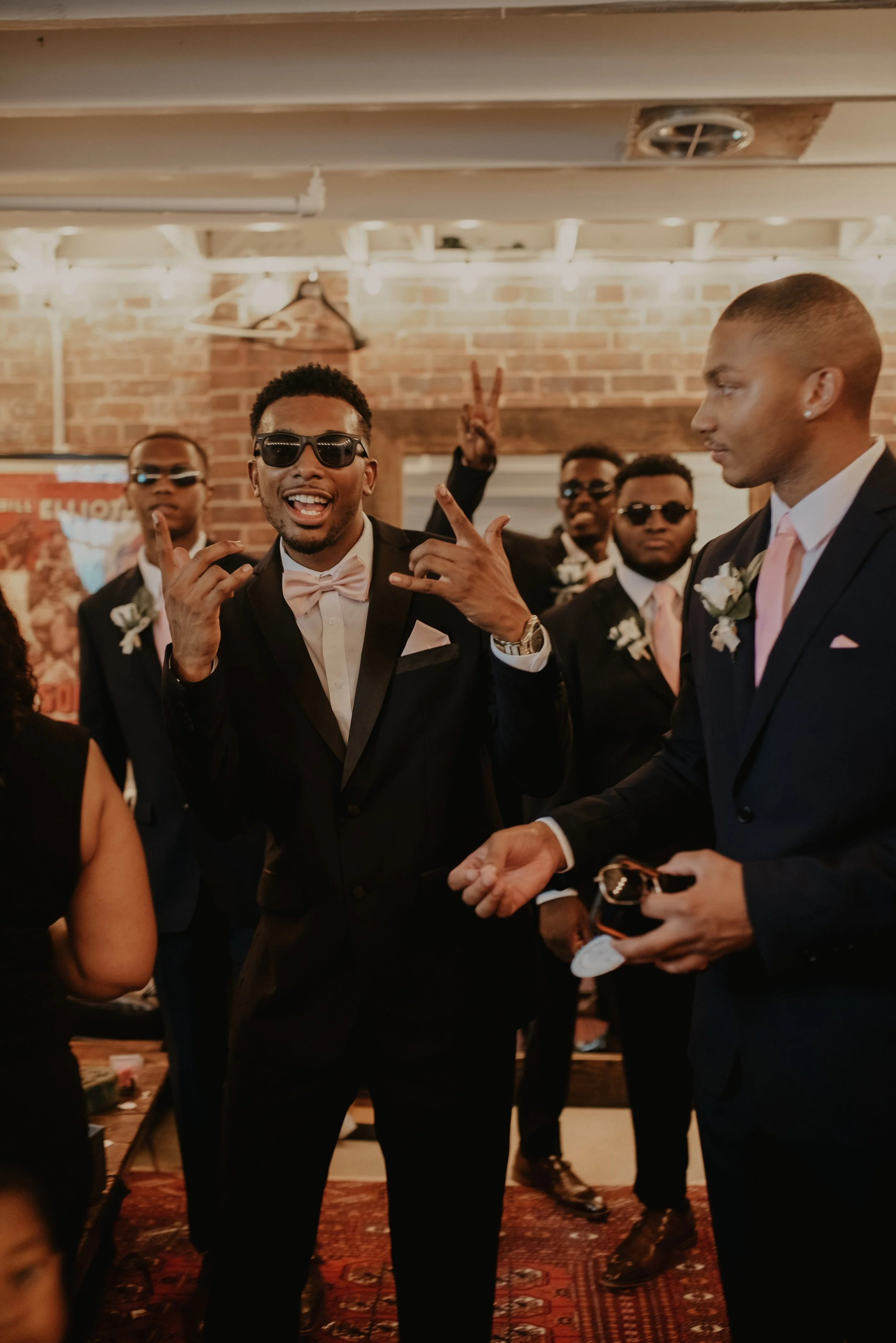 A group of men dressed in formal suits, some wearing sunglasses, at a celebration in a warmly lit room with brick walls. One man is making a hand gesture and smiling, while another man appears to be speaking or gesturing with his hands.
