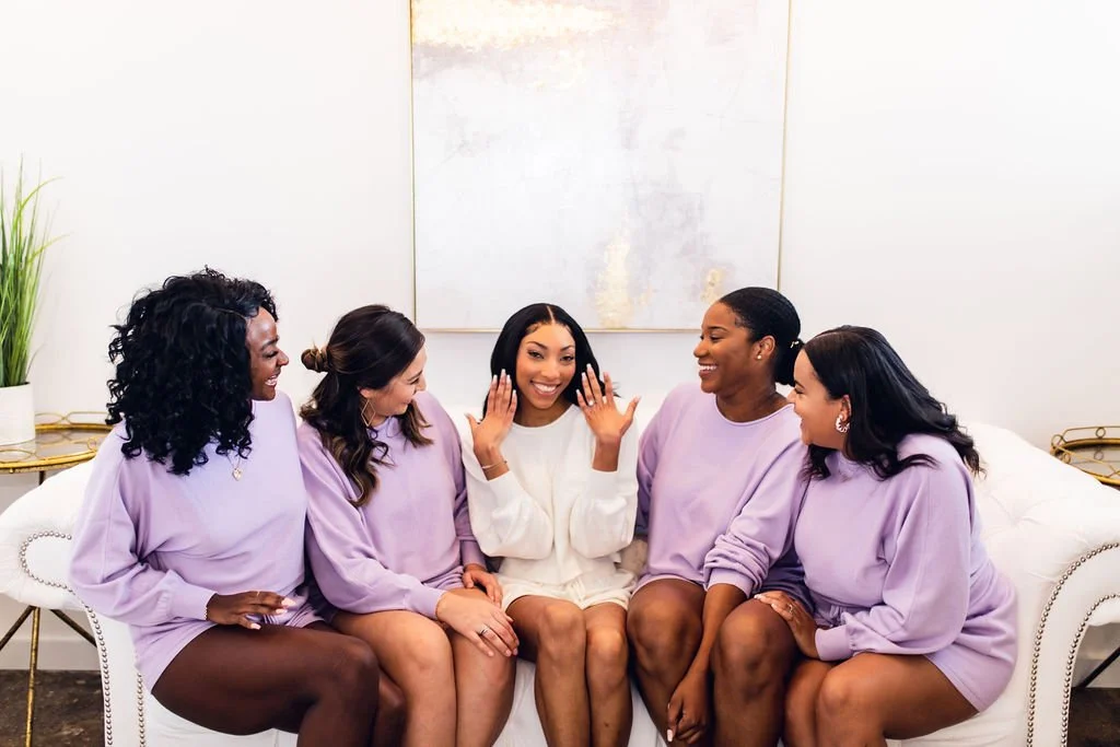 Five women sitting on a white sofa, smiling, with one woman in the center raising her hands, all dressed in light purple and cream outfits, in a bright, modern room.