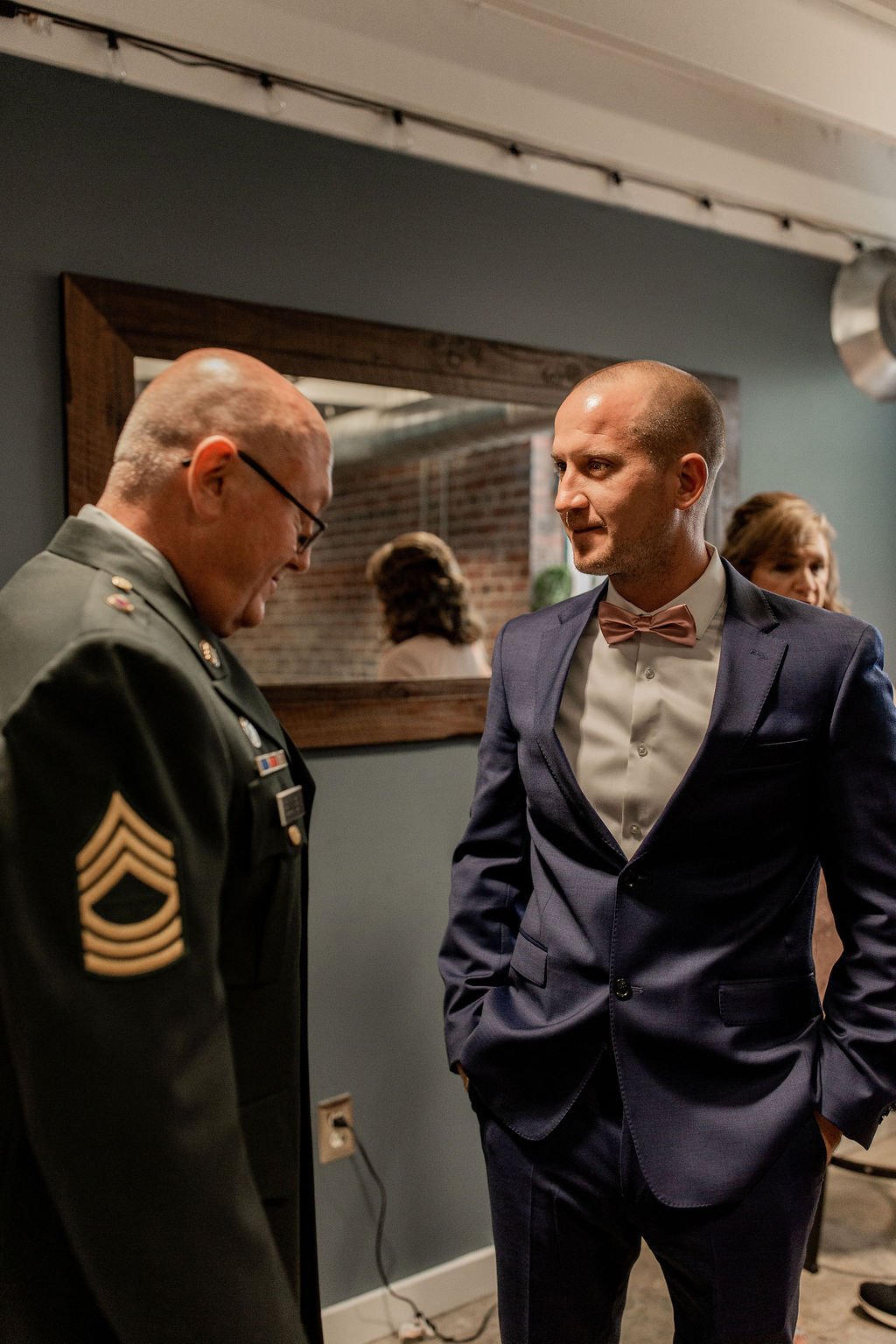 A man in a military uniform talking to a man in a dark tuxedo with a pink bow tie, indoors with a mirror and brick wall in the background.