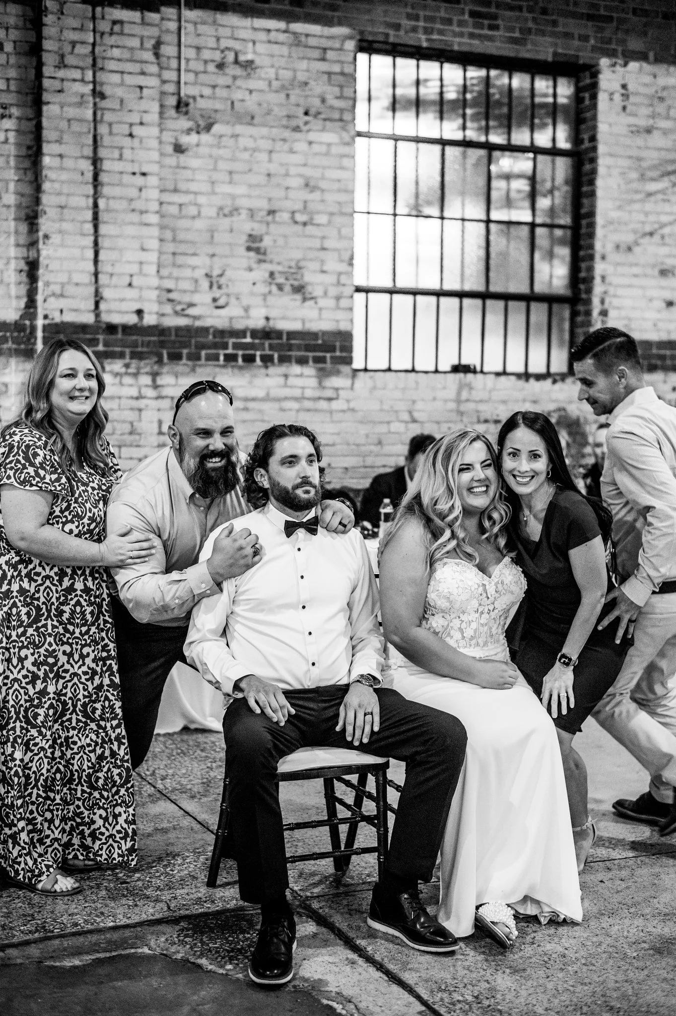 Black and white photo of six people at a wedding celebration, with a bride and groom sitting in the center surrounded by four friends, inside a brick-walled venue with large windows.