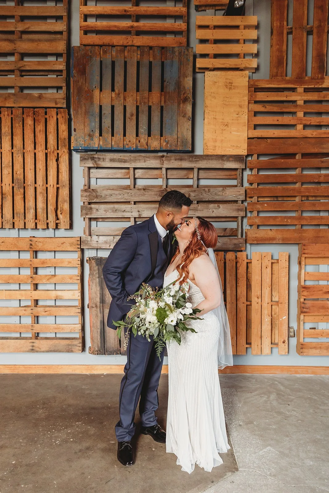A bride and groom sharing a kiss on their wedding day, standing in front of a rustic wood wall backdrop.