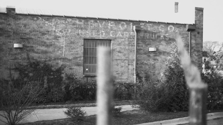 An old brick building with a faded sign on the wall, surrounded by bushes and a sidewalk, with blurred objects in the foreground.