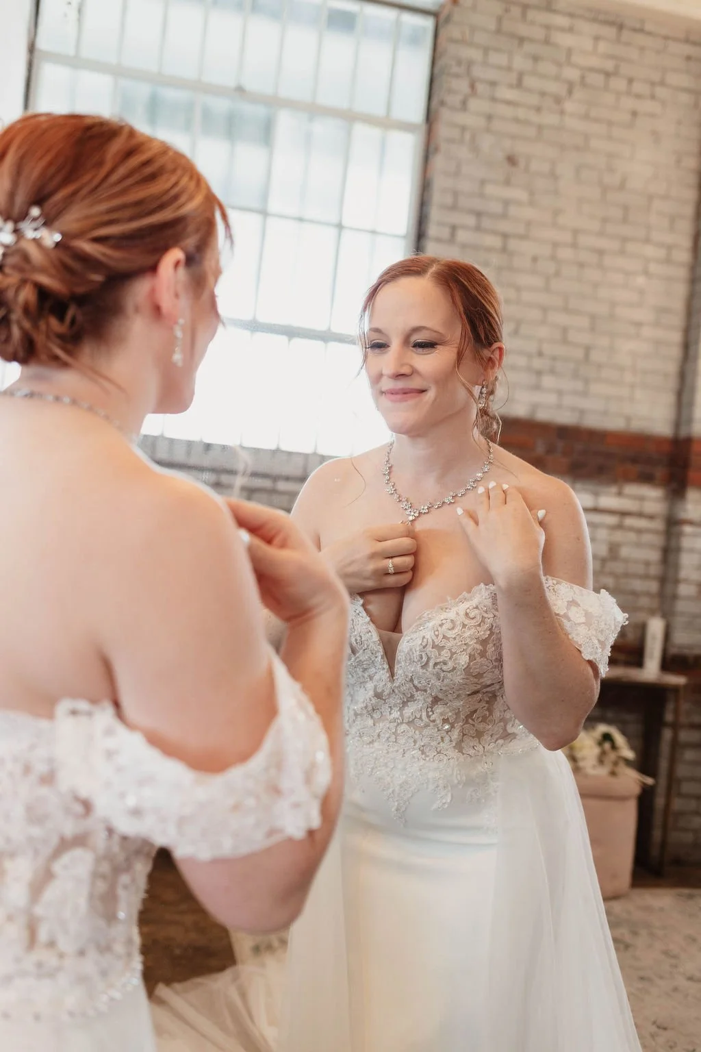 Two women in wedding dresses smiling and admiring each other, one touching her necklace, inside a rustic industrial venue with brick walls and large windows.