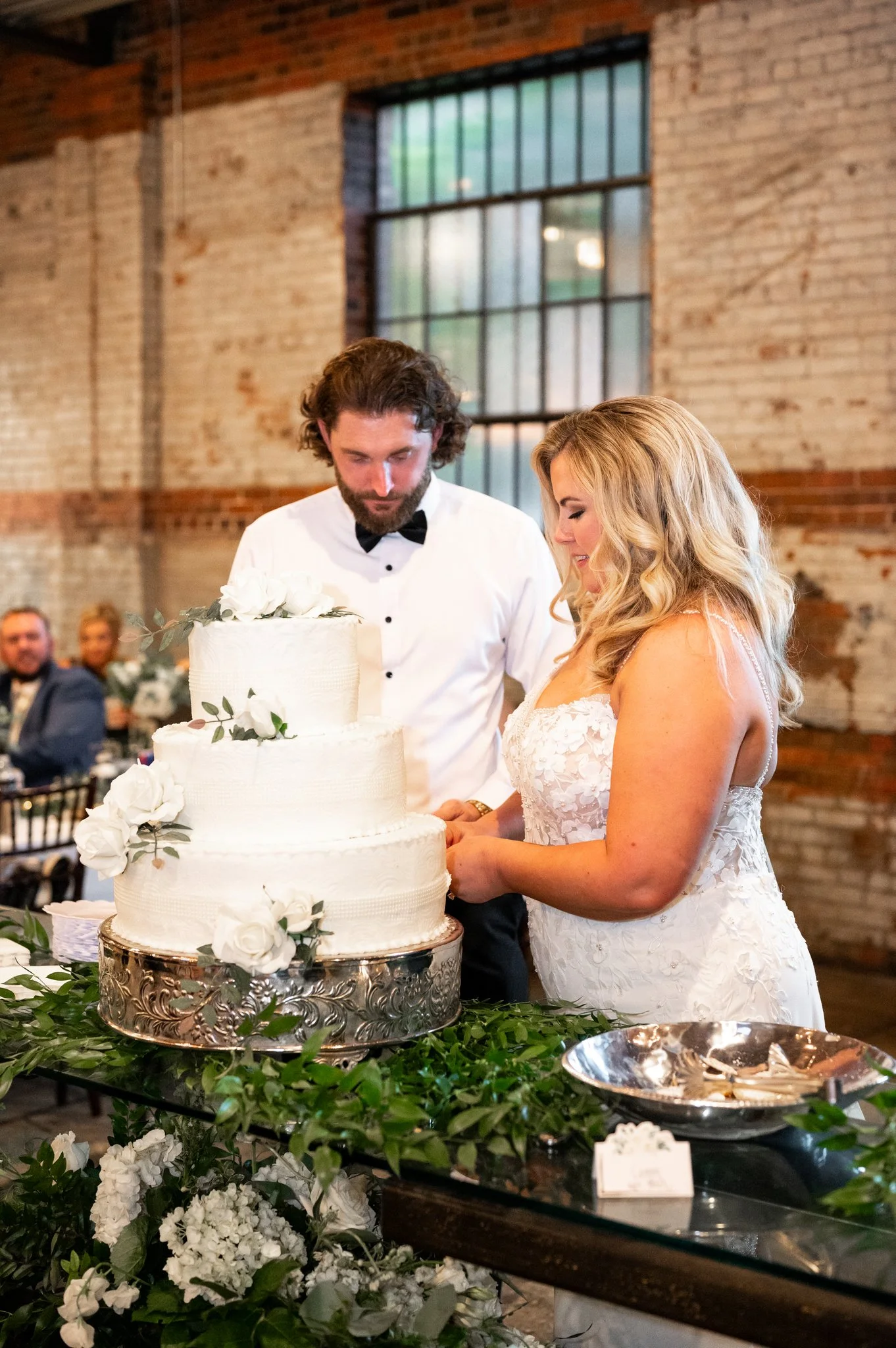 A bride and groom cutting a wedding cake decorated with white flowers and green leaves at their wedding reception in a rustic venue with exposed brick walls and large windows.