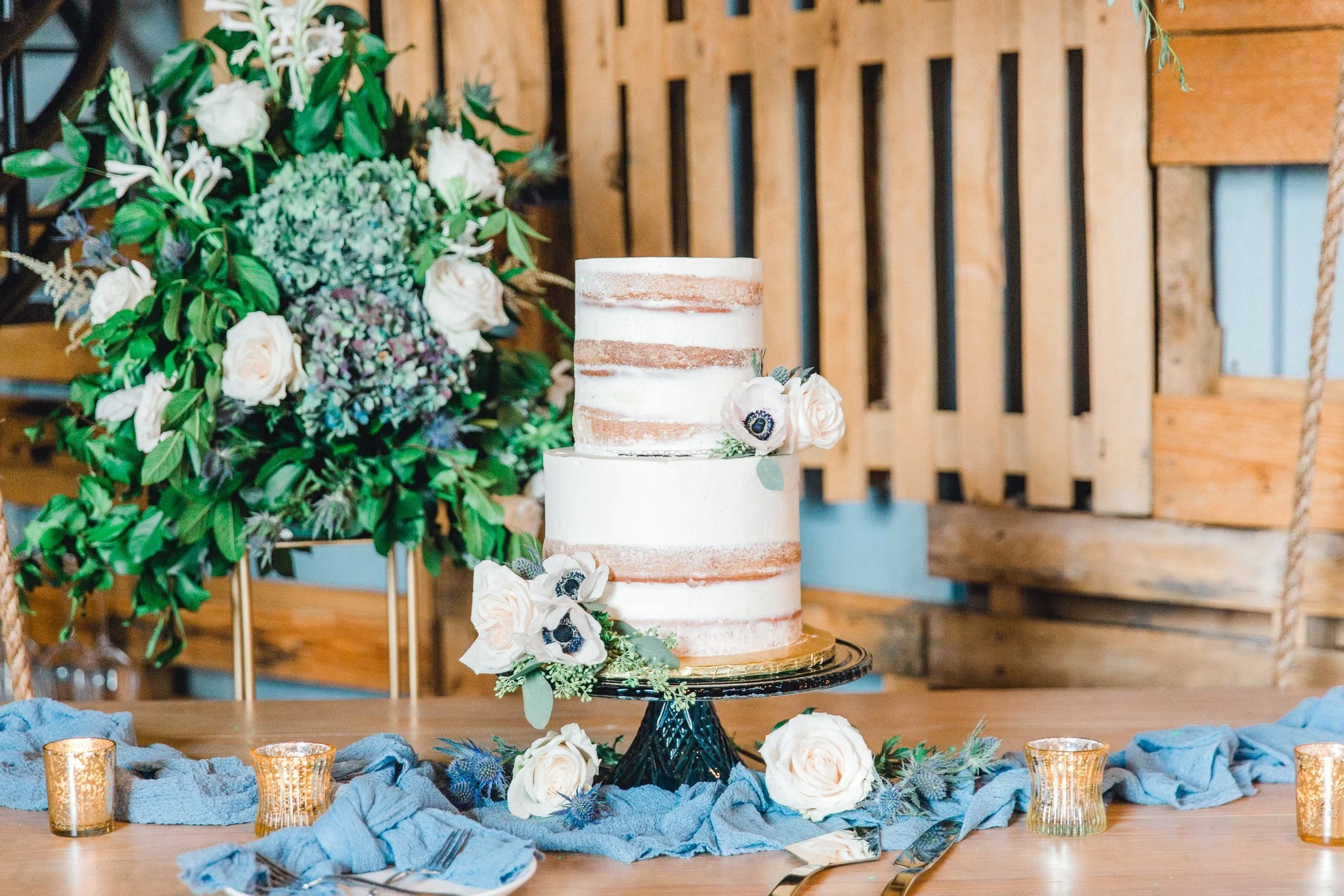 A two-tiered semi-naked wedding cake decorated with white flowers and greenery on a black cake stand, surrounded by blue fabric and small gold candle holders, set on a wooden table with a large floral arrangement in the background.