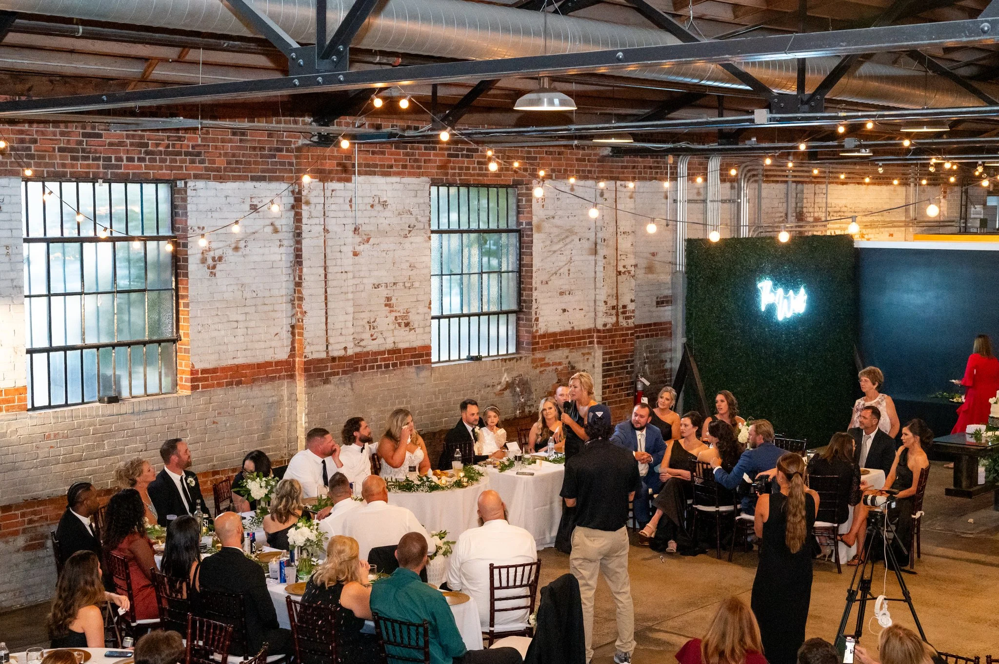 A wedding reception in an industrial-style venue with brick walls, large windows, hanging string lights, and a group of people sitting at a head table listening to a speech.