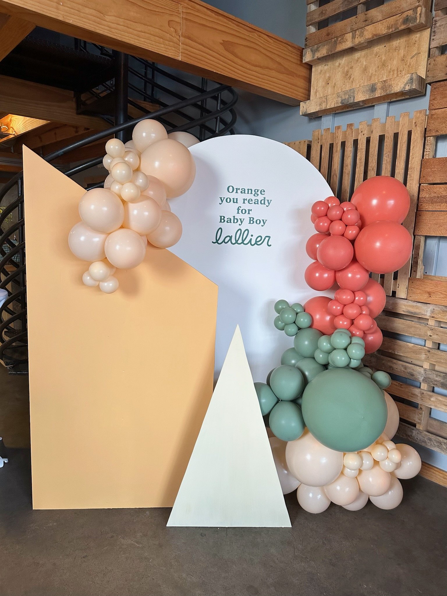 Decorative setup with pastel-colored balloons in shades of peach, pink, and green arranged around a white circular backdrop. The backdrop has text that says, 'Orange you ready for Baby Boy fallier.' There are also large geometric panels in peach, white, and cream to complement the balloon arrangement.