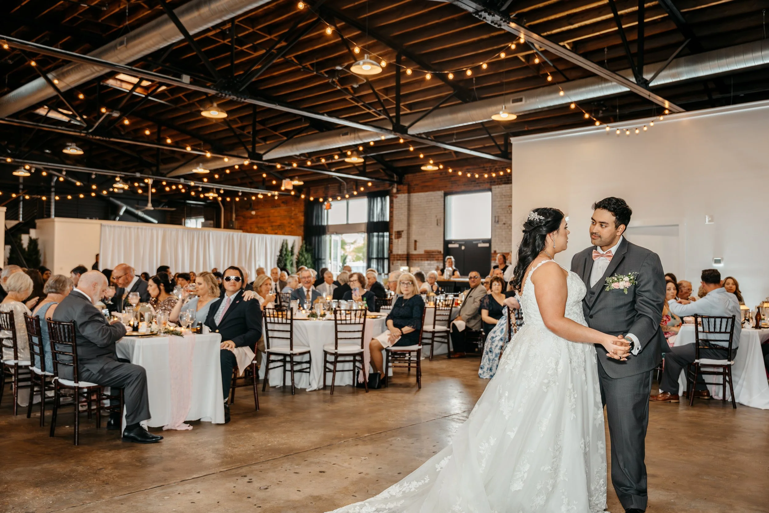 A bride and groom dancing at their wedding reception in a spacious industrial-style venue with string lights and many seated guests watching at Studio 215 a modern warehouse venue in Fayetteville, NC