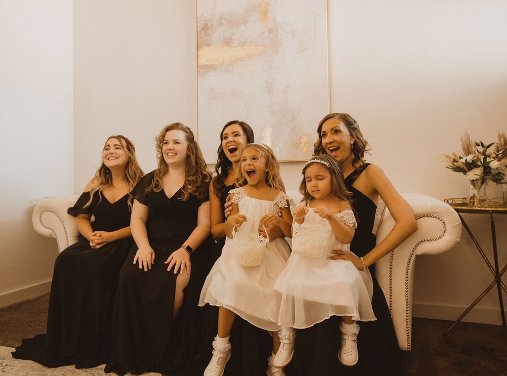 Six women and girls dressed in black and white, sitting on a white couch and laughing, in a room with beige walls and floral decor.