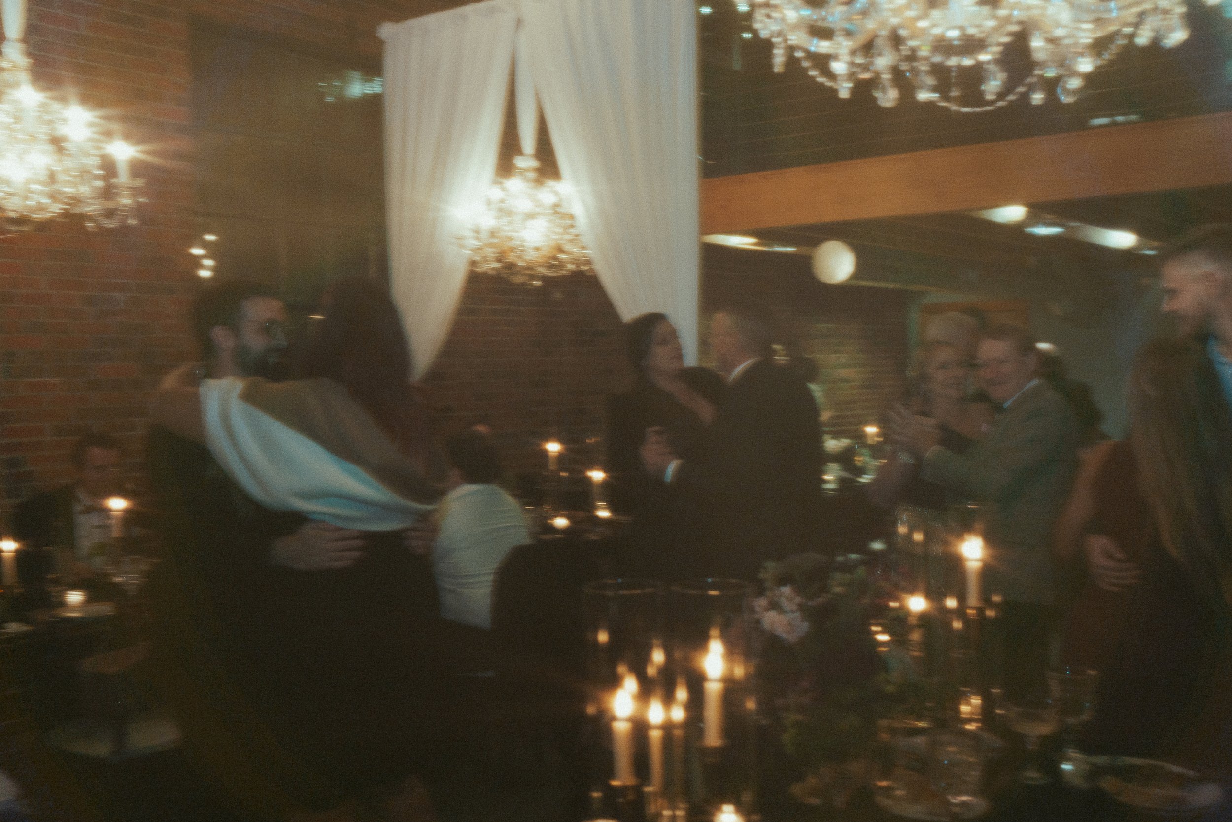 People dancing and socializing at a dimly lit wedding reception or party with chandeliers and candles.