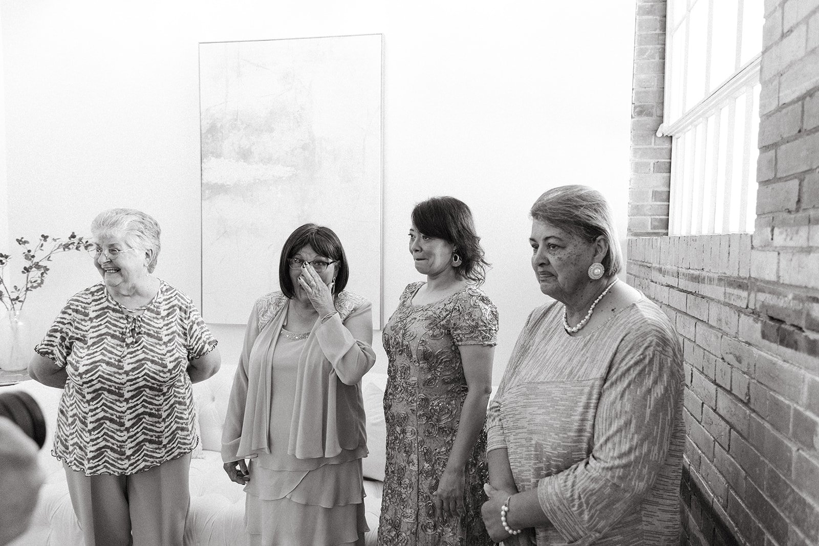 Four women standing in a room with a white wall, a window, and a brick wall, some smiling and one wiping her eye.