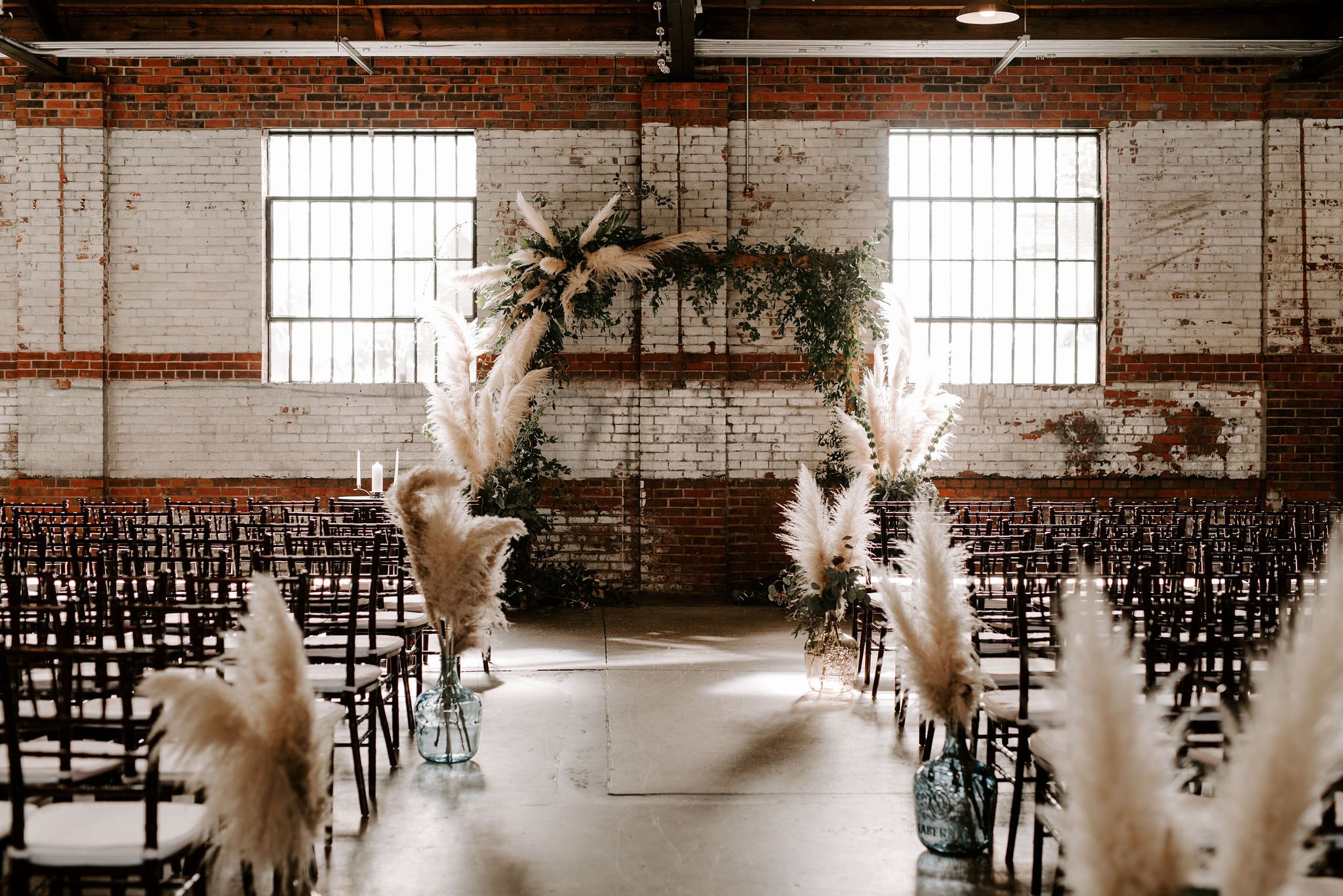 An indoor event space with exposed brick walls and large industrial windows, decorated with tall vases of pampas grass and greenery forming a wedding or celebration arch, with rows of chairs on either side.