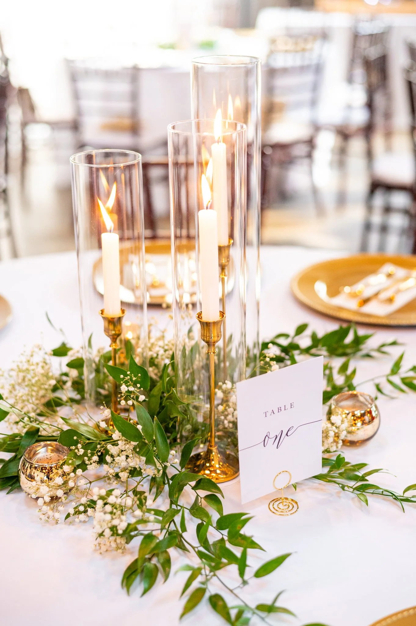Elegant table centerpiece with three tall glass candleholders containing white candles with lit flames, surrounded by greenery, small white flowers, and gold accents, on a white tablecloth at a formal event.