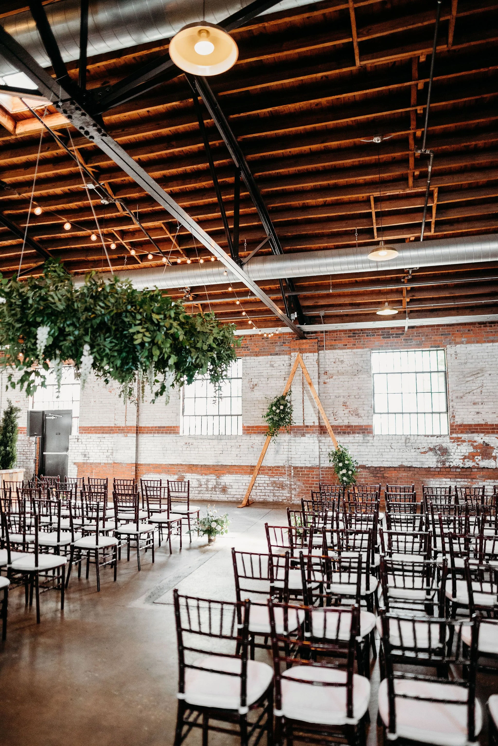 Wedding ceremony setup in an industrial-style venue with rows of black chairs, a wooden arch decorated with greenery and flowers, hanging greenery with lights, and brick walls with large windows.