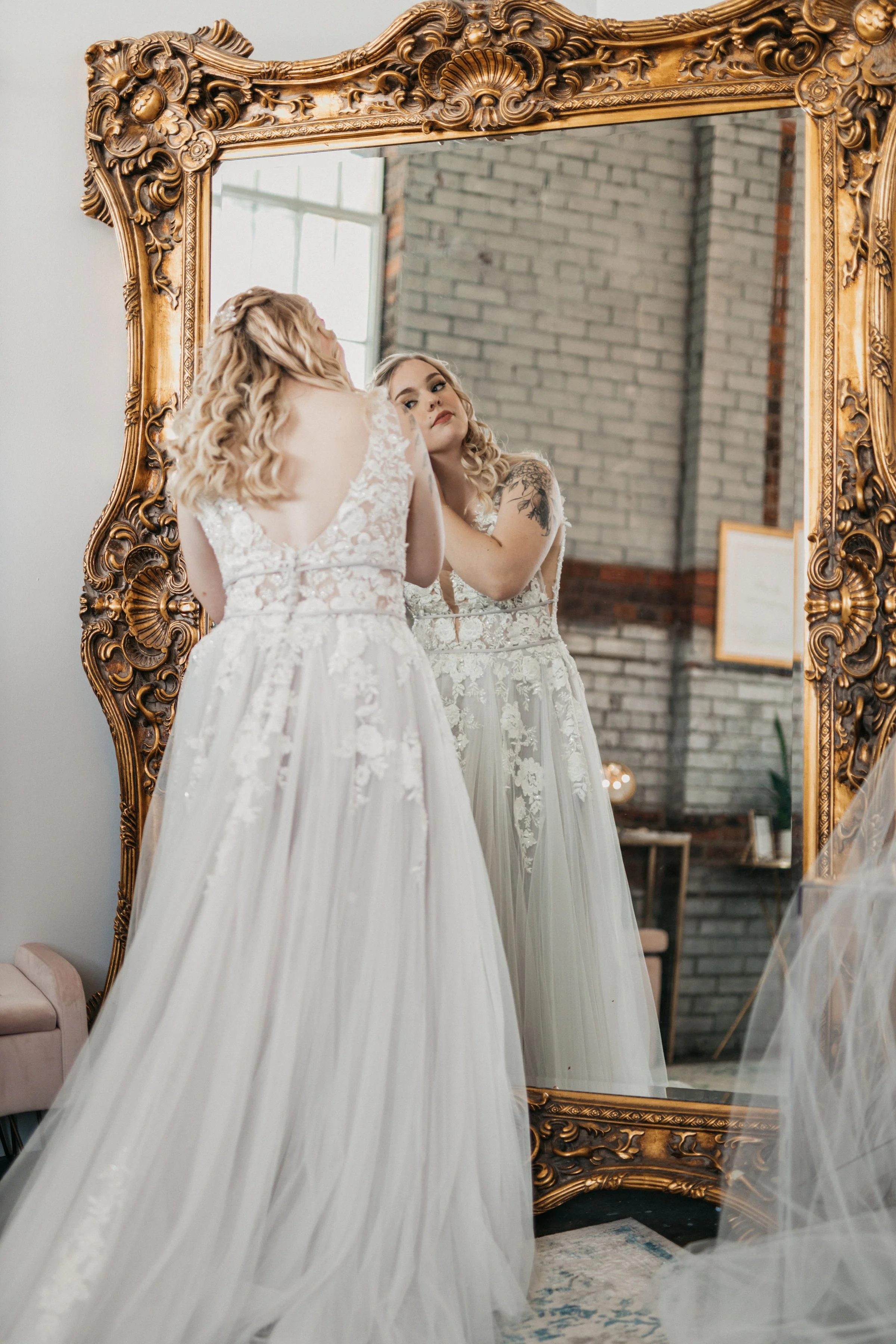 A woman in a wedding dress is looking at herself in an ornate gold-framed mirror, adjusting her hairstyle.