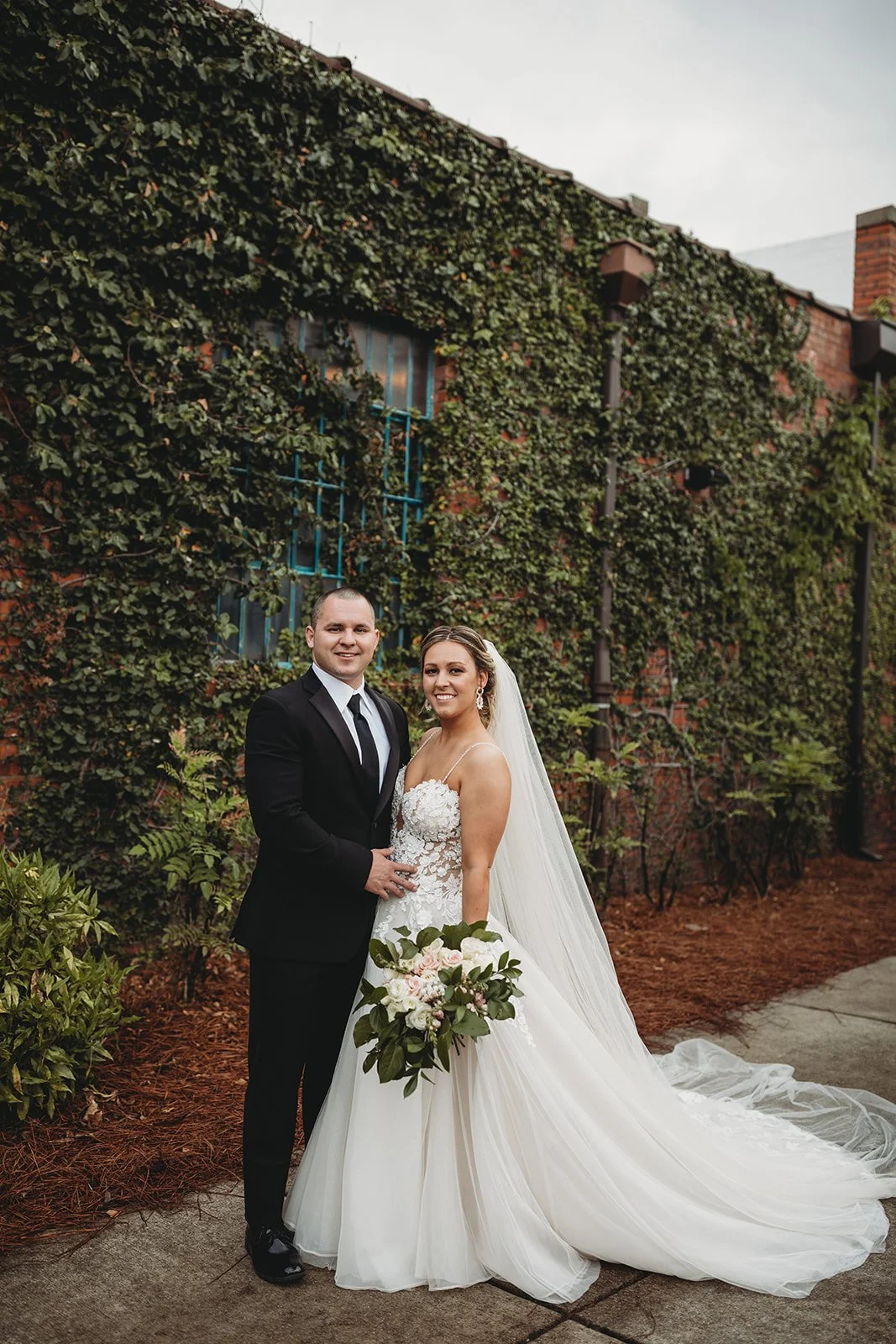 Bride and groom posing in front of the signature green ivy wall at Studio 215 in downtown Fayetteville, NC; a lush urban garden photography backdrop at our industrial warehouse venue near Fort Bragg and Raleigh.