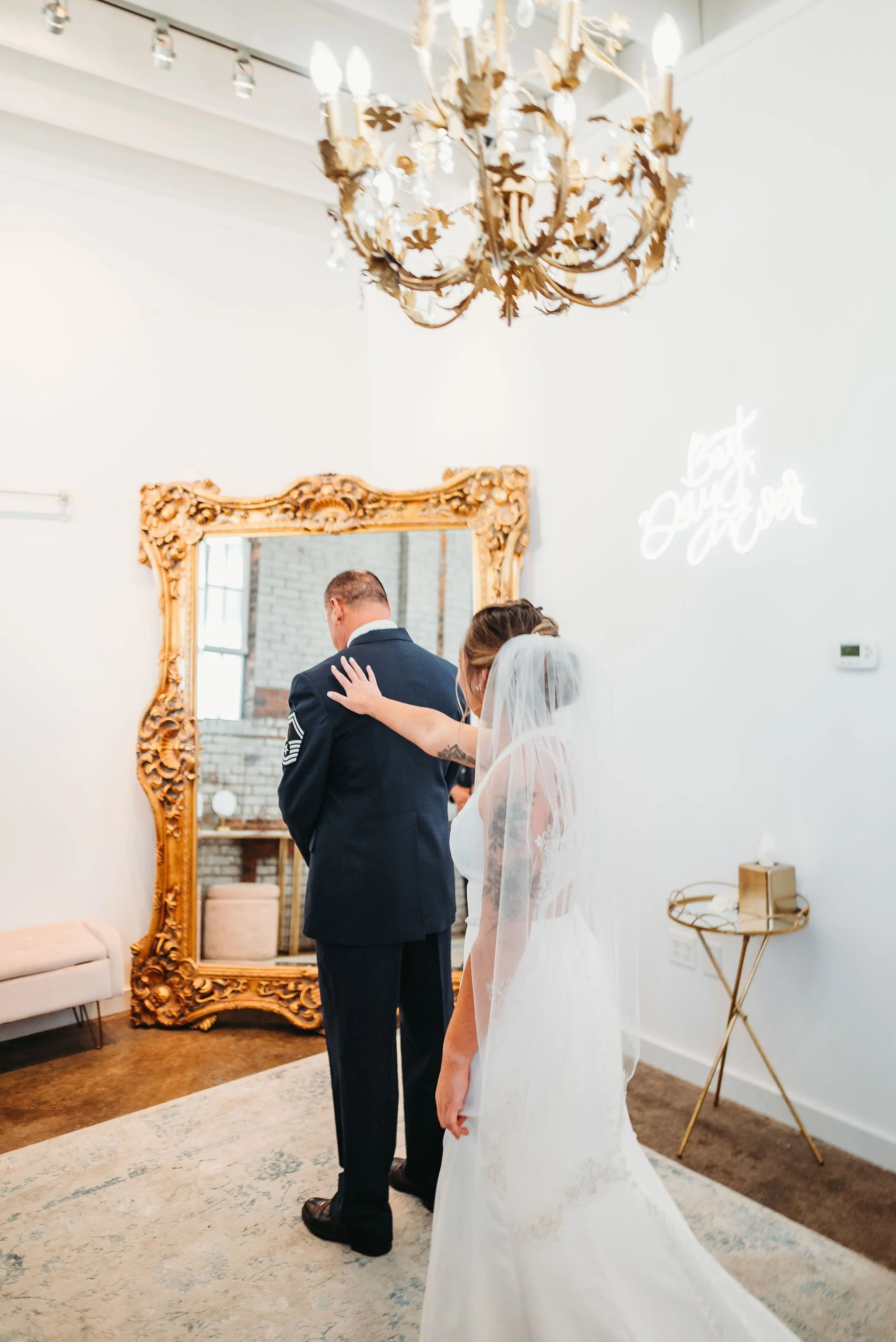 A bride in a white wedding dress and veil touches a groom in a military uniform during a wedding ceremony inside a decorated room with a large ornate mirror and a chandelier.