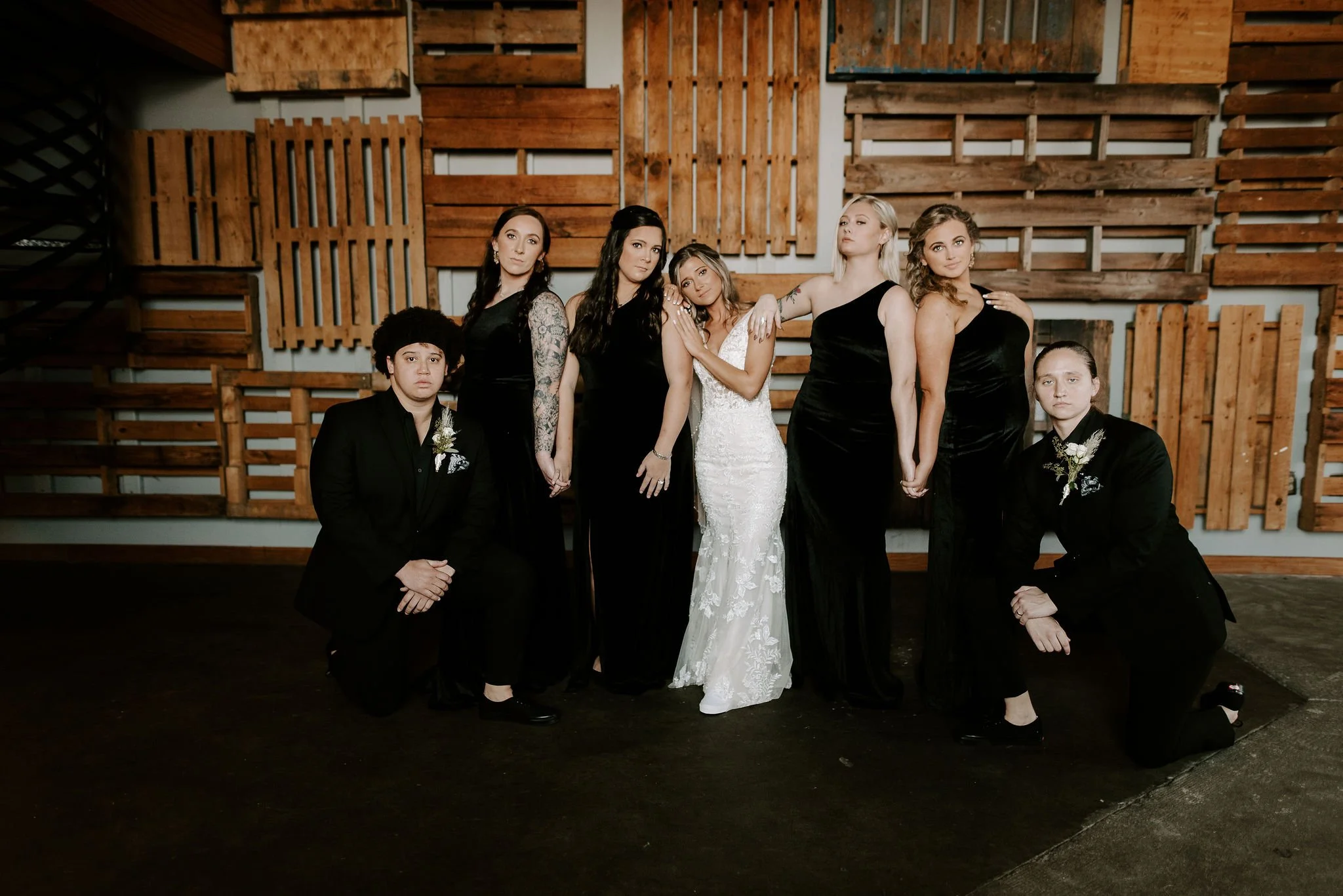 A group of eight people, including a bride in a white wedding dress and seven women in black dresses, pose together indoors in front of a wooden wall. Two of the women are kneeling. The setting appears to be a wedding or formal event.