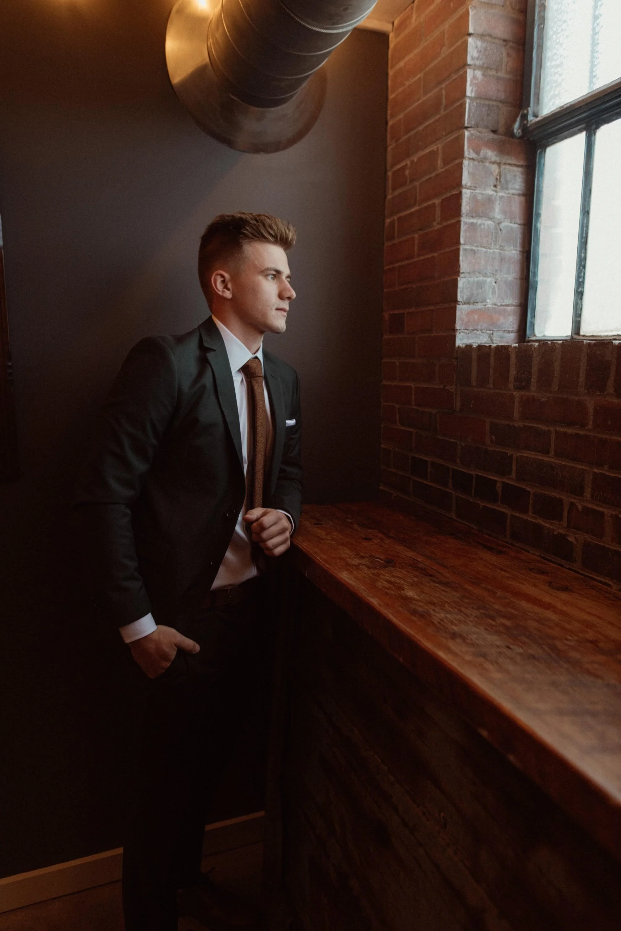 A young man in a black suit, white shirt, and tie stands indoors near a wooden countertop, looking out a window on his right in a dimly lit room with exposed brick walls and a large metal pipe overhead.