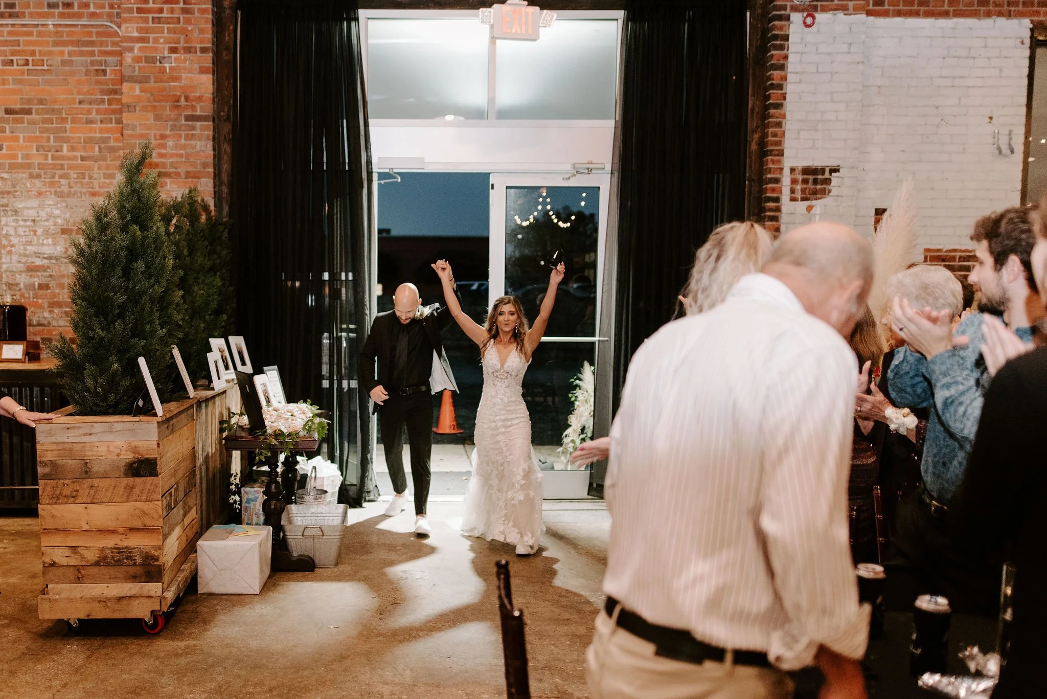 Bride and groom entering reception area, guests clapping, rustic decor with brick walls, black curtains, and a potted plant.