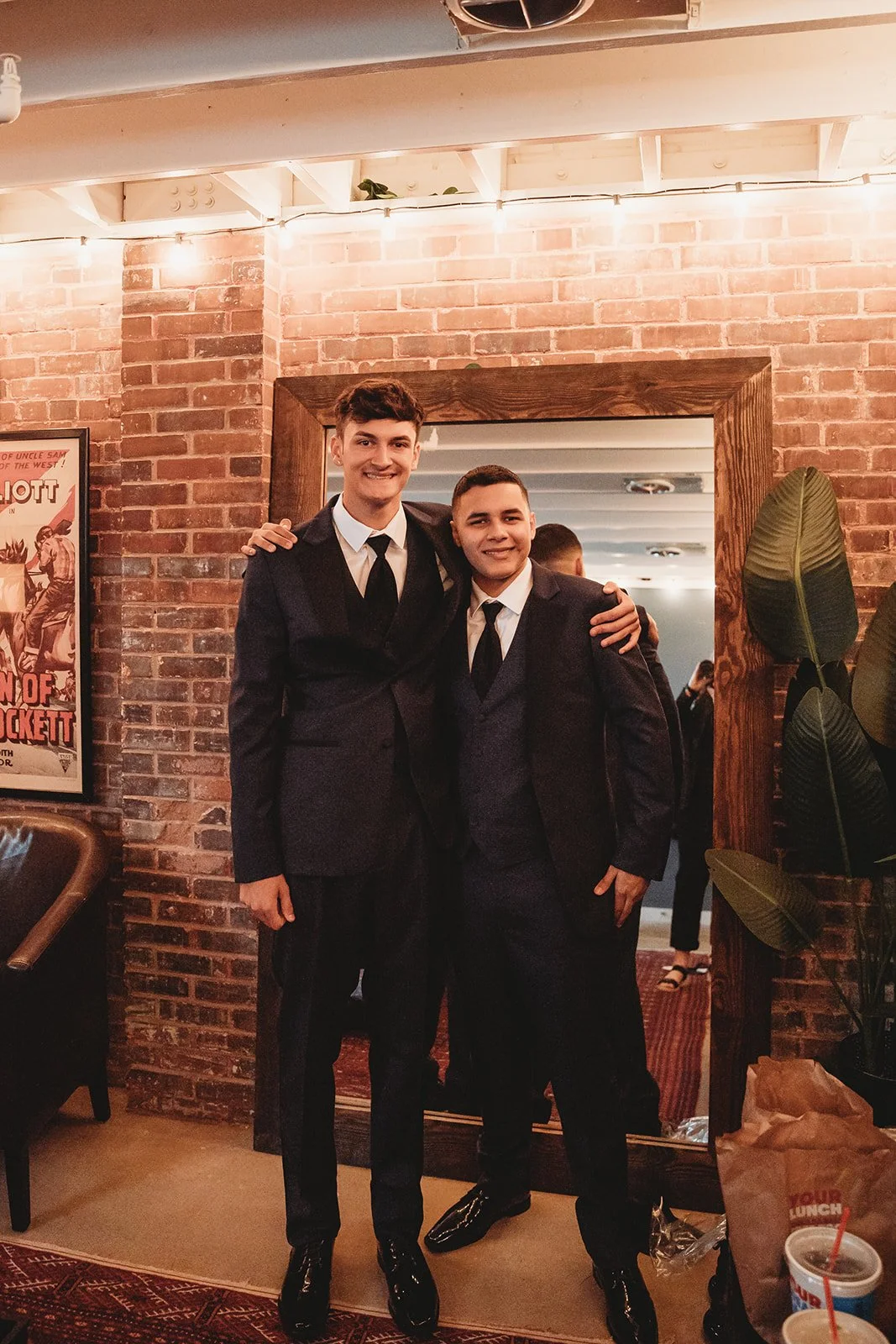 Two young men in formal suits and ties smiling and posing together in front of a mirror in a brick-walled room, possibly a restaurant or bar.