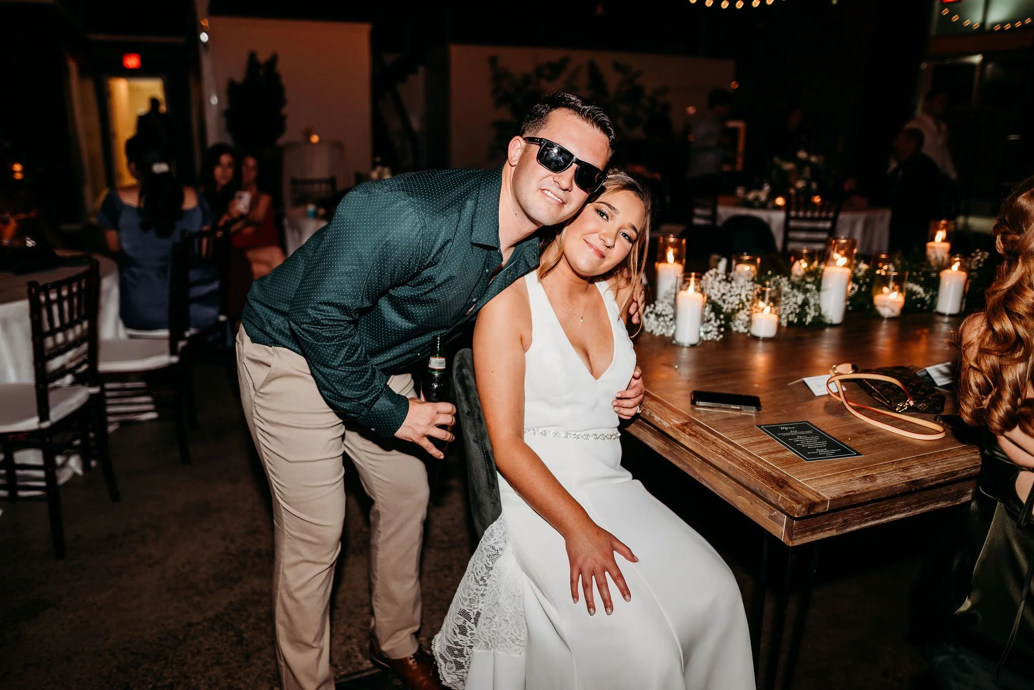 A man and woman at a dimly lit indoor social event or wedding reception. The man is wearing sunglasses, a dark long-sleeve shirt, and khaki pants, and is holding a drink. The woman is seated, wearing a white dress, and smiling. They are leaning towar