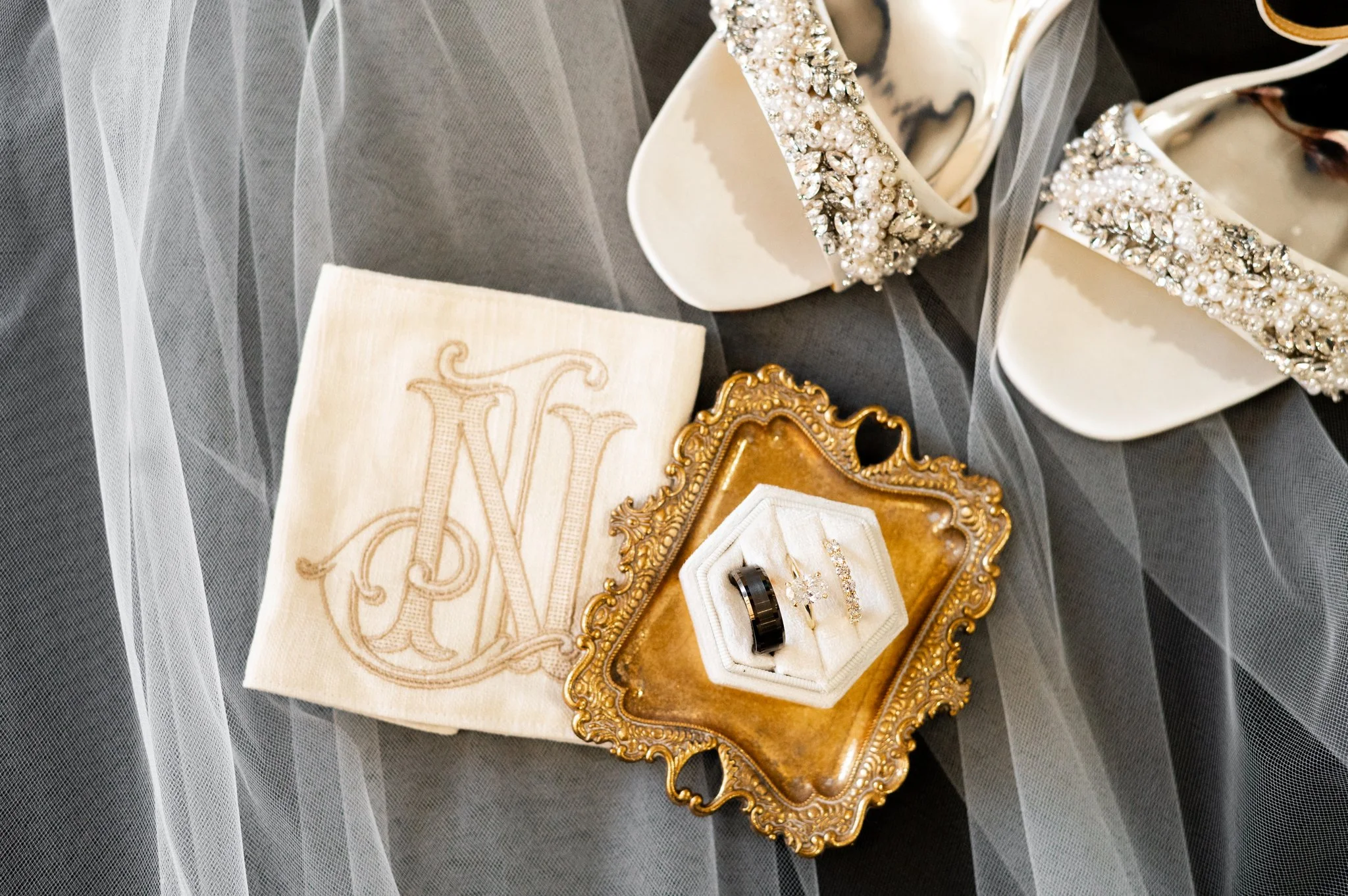 Elegant vintage wedding accessories including a monogrammed cloth, pearl and rhinestone embellished shoes, a gold ornate jewelry tray with a black wedding band and diamond jewelry, and a sheer black veil in the background.