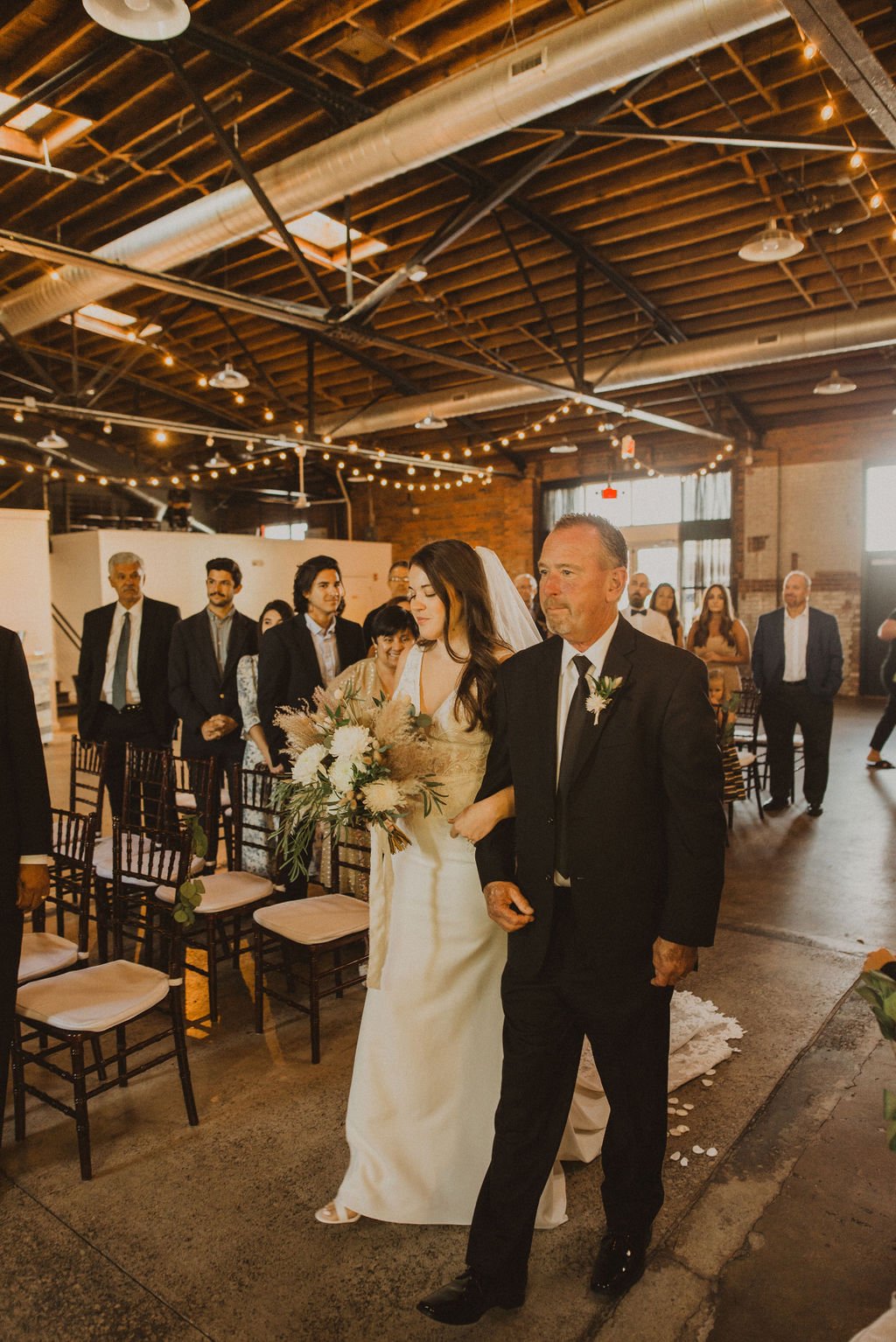 A bride in a white wedding gown holding a bouquet of flowers is walking down the aisle, accompanied by an older man dressed in a black suit, at a wedding ceremony in a rustic indoor venue with string lights and exposed wooden beams.