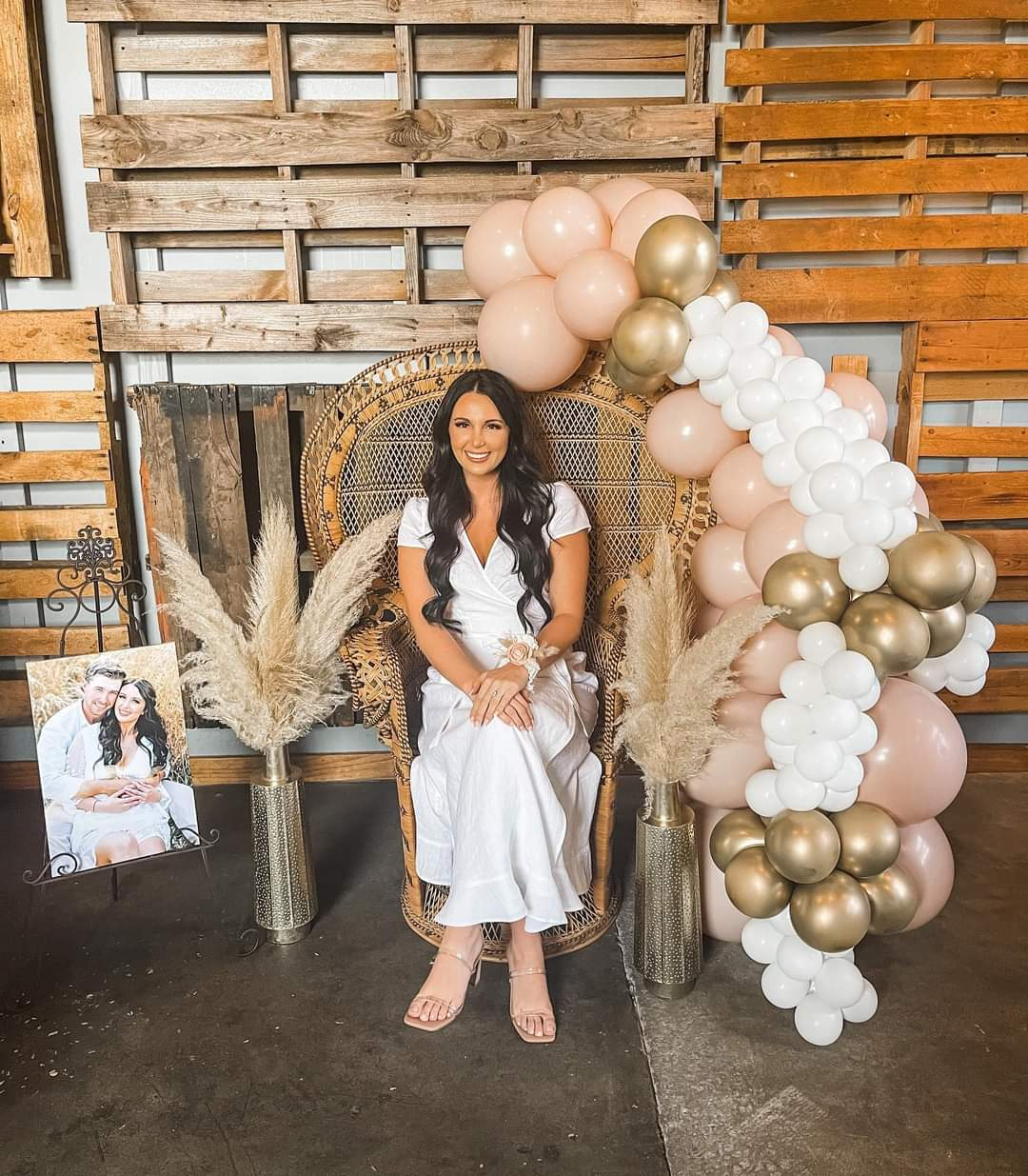 A woman in a white dress sitting on a decorative peacock chair is surrounded by pink, white, and gold balloons and pampas grass in vases, with a wooden pallet backdrop and a framed photo on an easel.