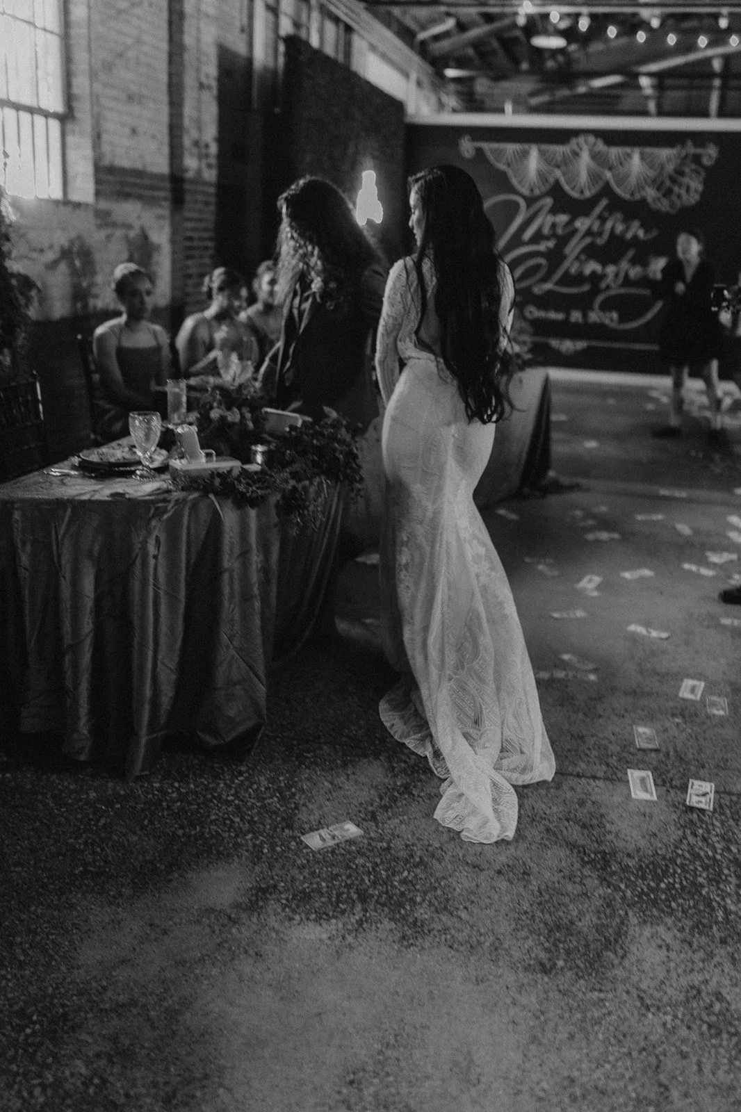 Black and white photo of a woman in a white dress at a wedding reception, standing near a table with guests seated. Money is scattered on the floor around her. A decorated wall with a sign that reads 'Wedding Lindsay' is in the background.