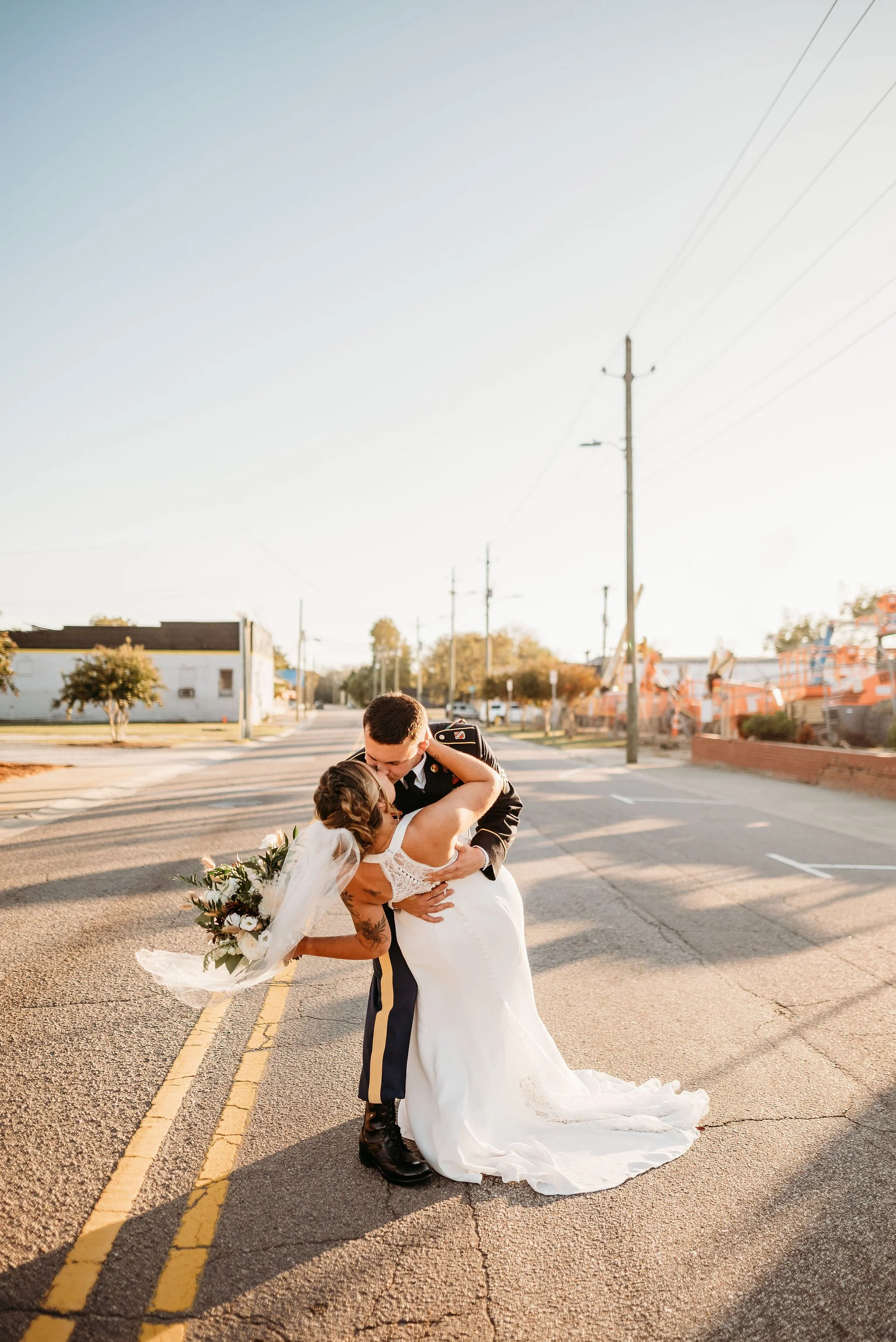 A newlywed couple sharing a kiss on an empty city street at sunset, the groom in military uniform and the bride holding a bouquet, both dressed in wedding attire.