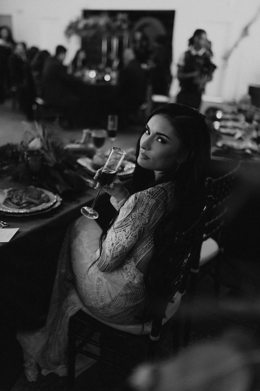 A woman with long dark hair and makeup, wearing an intricately patterned dress, sitting at a dining table holding a glass of red wine in a black-and-white photo at a social gathering.