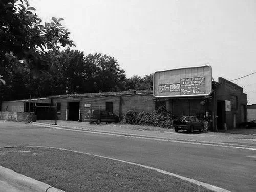 A black and white photo of a single-story building with an outdoor billboard sign, parked cars, and trees in the background.