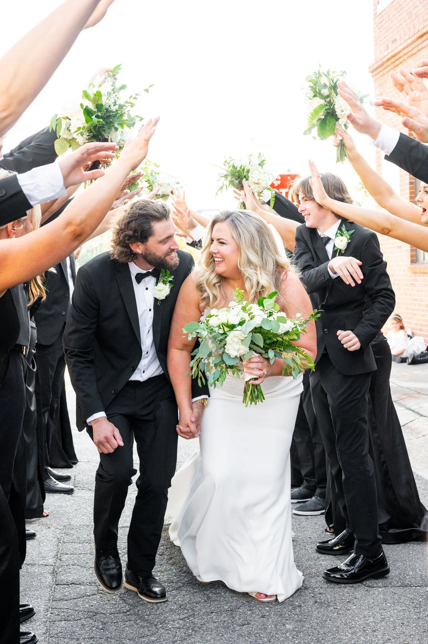 A wedding celebration with a bride and groom walking through a crowd of friends and family, holding hands, with everyone raising bouquets and cheering.