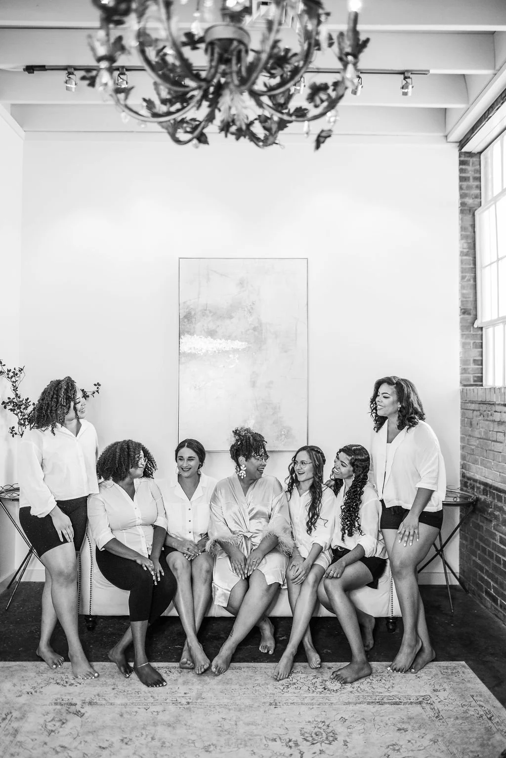 A group of seven women sitting and standing in a living room, smiling and interacting with each other. They are dressed casually, some in shirts and shorts, and one in a satin robe. The room has minimalist decor with a large painting on the wall, a c