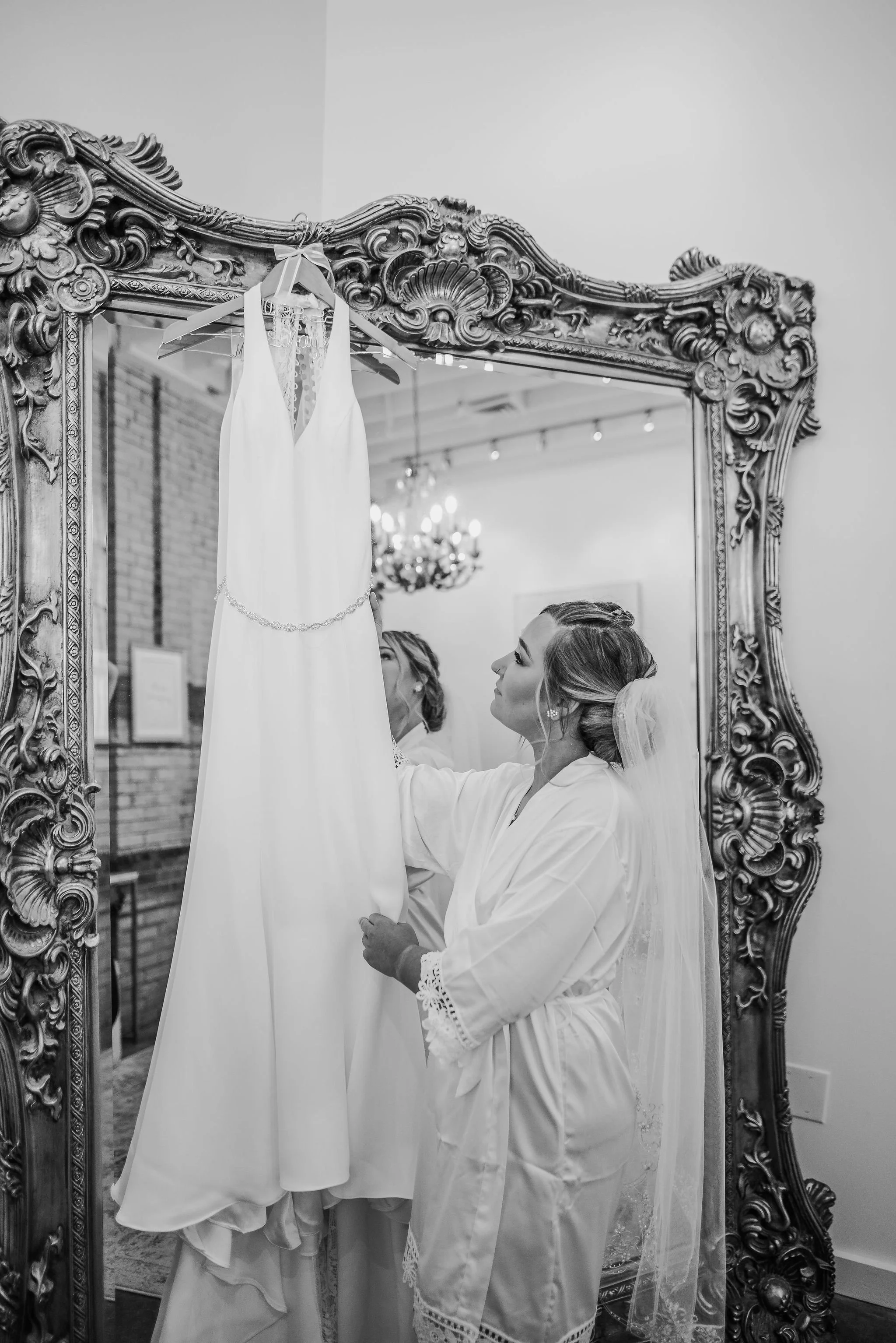 A woman in a satin robe and veil looks at a wedding dress hanging on a hanger in an ornate mirror.