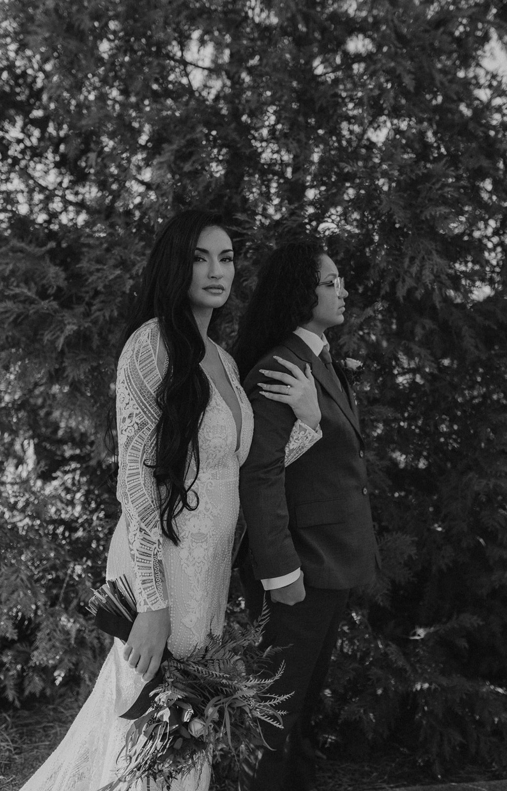 A black and white photo of two women standing outdoors with trees in the background. One woman in a lace wedding dress holds a bouquet and looks at the camera, while the other woman in a suit and glasses stands behind her with her hand on her shoulde