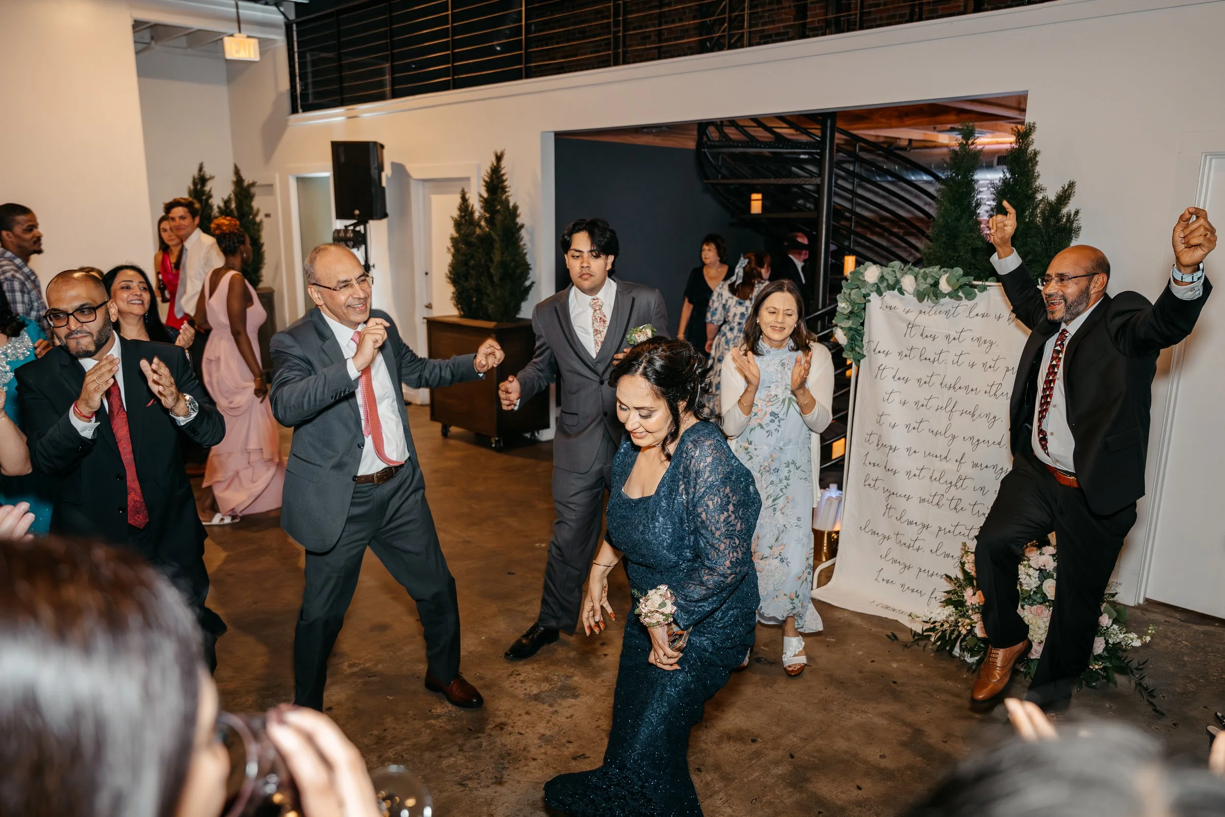 Guests dancing and celebrating at a wedding reception with a woman in a dark blue lace dress in the center, surrounded by other guests in formal attire, with a decorative backdrop and potted plants nearby.