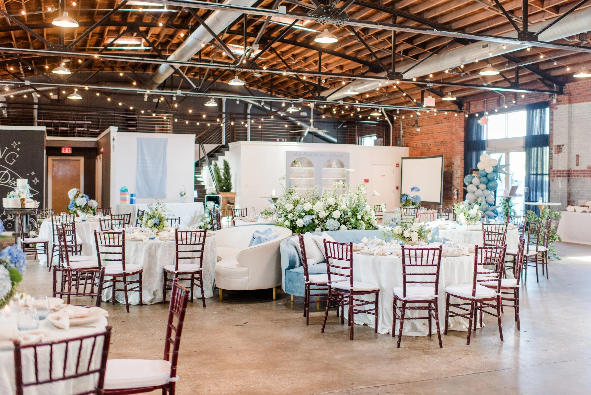 Wedding reception setup in a loft-style venue with round tables covered in white tablecloths, decorated with white and blue flower centerpieces, surrounded by wooden chairs, and a large floral arrangement in the center. String lights and balloons are