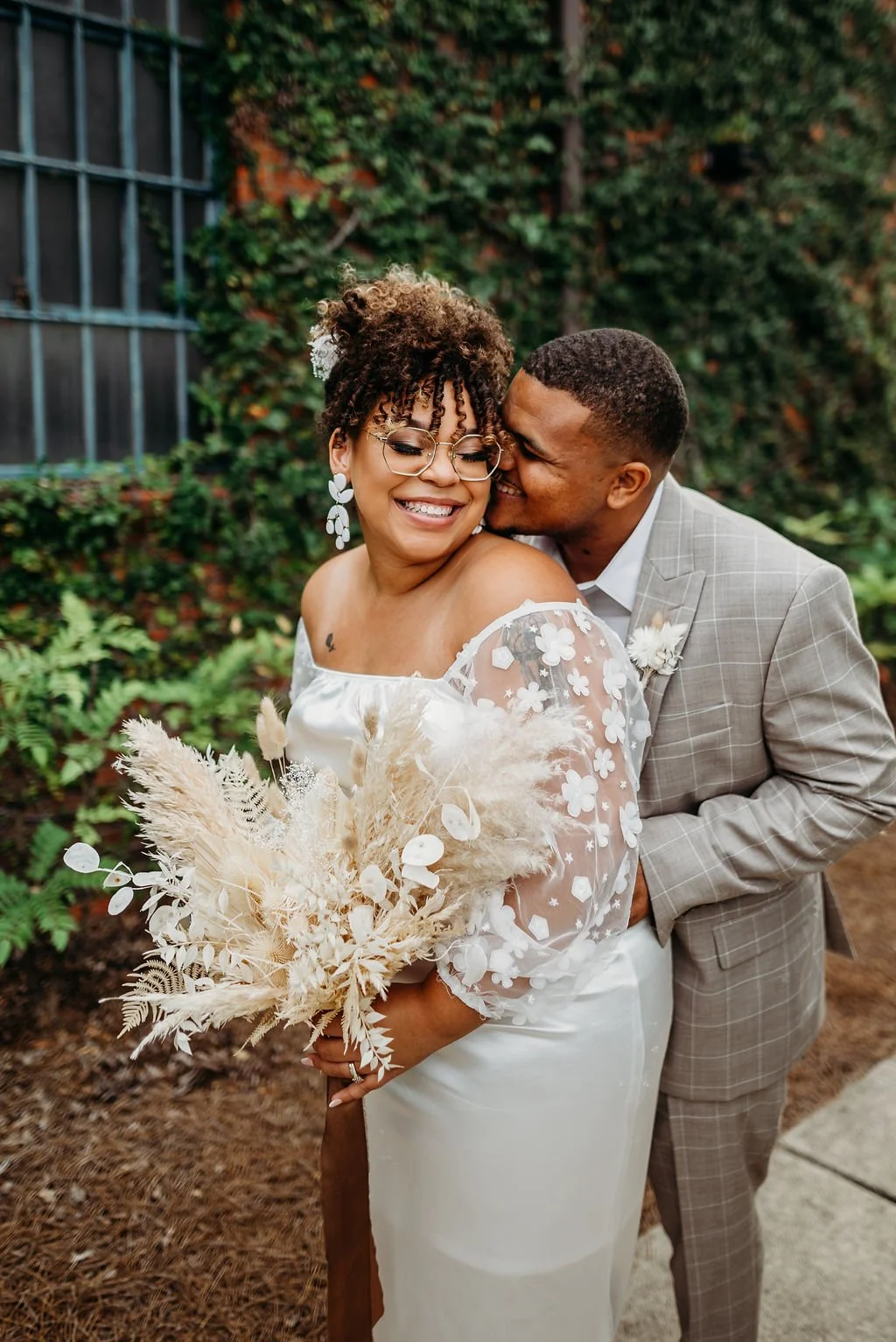 A joyful couple dressed in wedding attire, embracing outdoors with greenery in the background. The woman holds a bouquet of dried flowers, and they are sharing a moment of happiness.