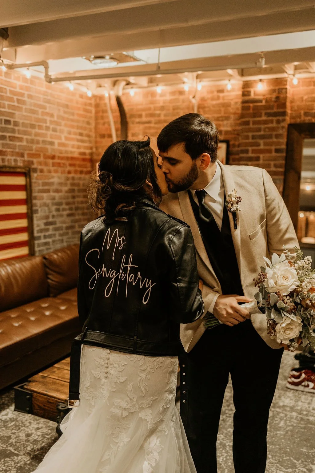 A bride and groom share a kiss in a cozy, brick-walled room decorated with string lights. The bride wears a leather jacket with 'Mrs Sngletary' written on the back and a white wedding gown. The groom wears a light-colored suit jacket and holds a bouquet of white flowers.