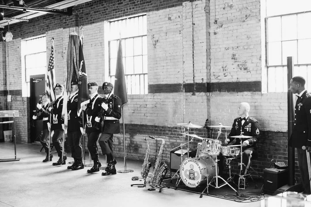 Military band in uniform standing in a line, holding flags and instruments inside a brick-walled room with large windows.