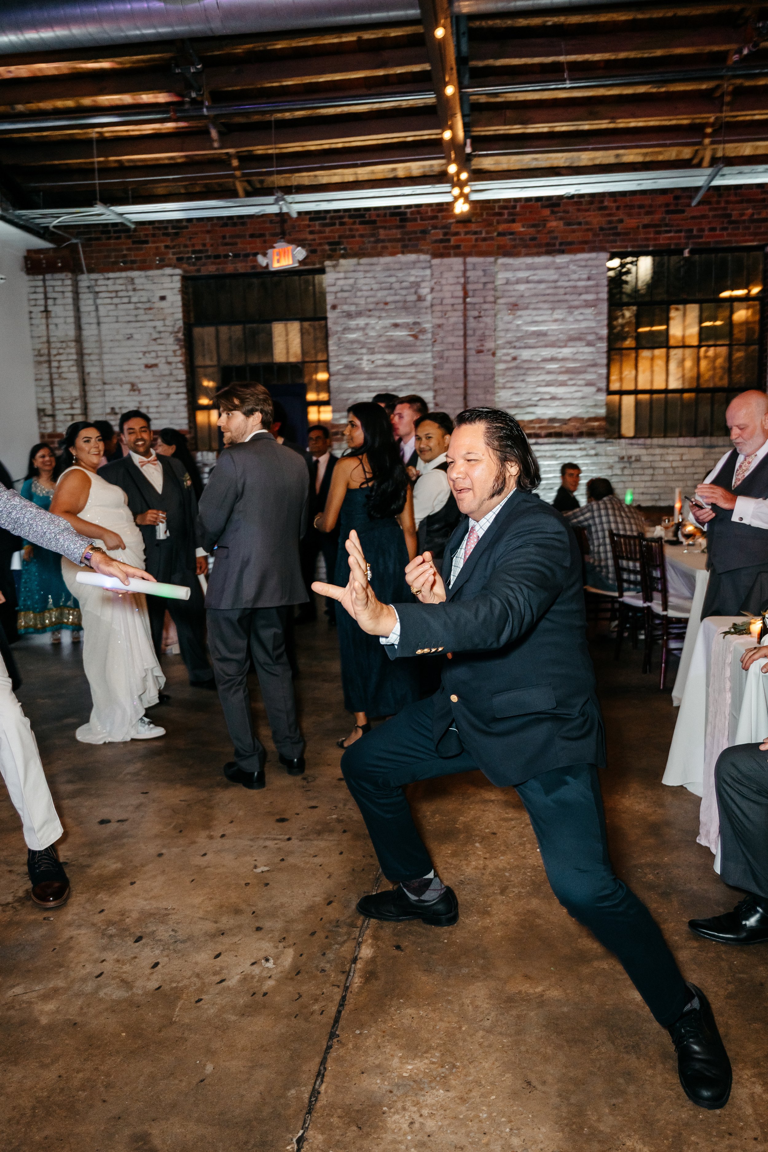 A man in a suit dancing at a wedding reception with guests in the background.
