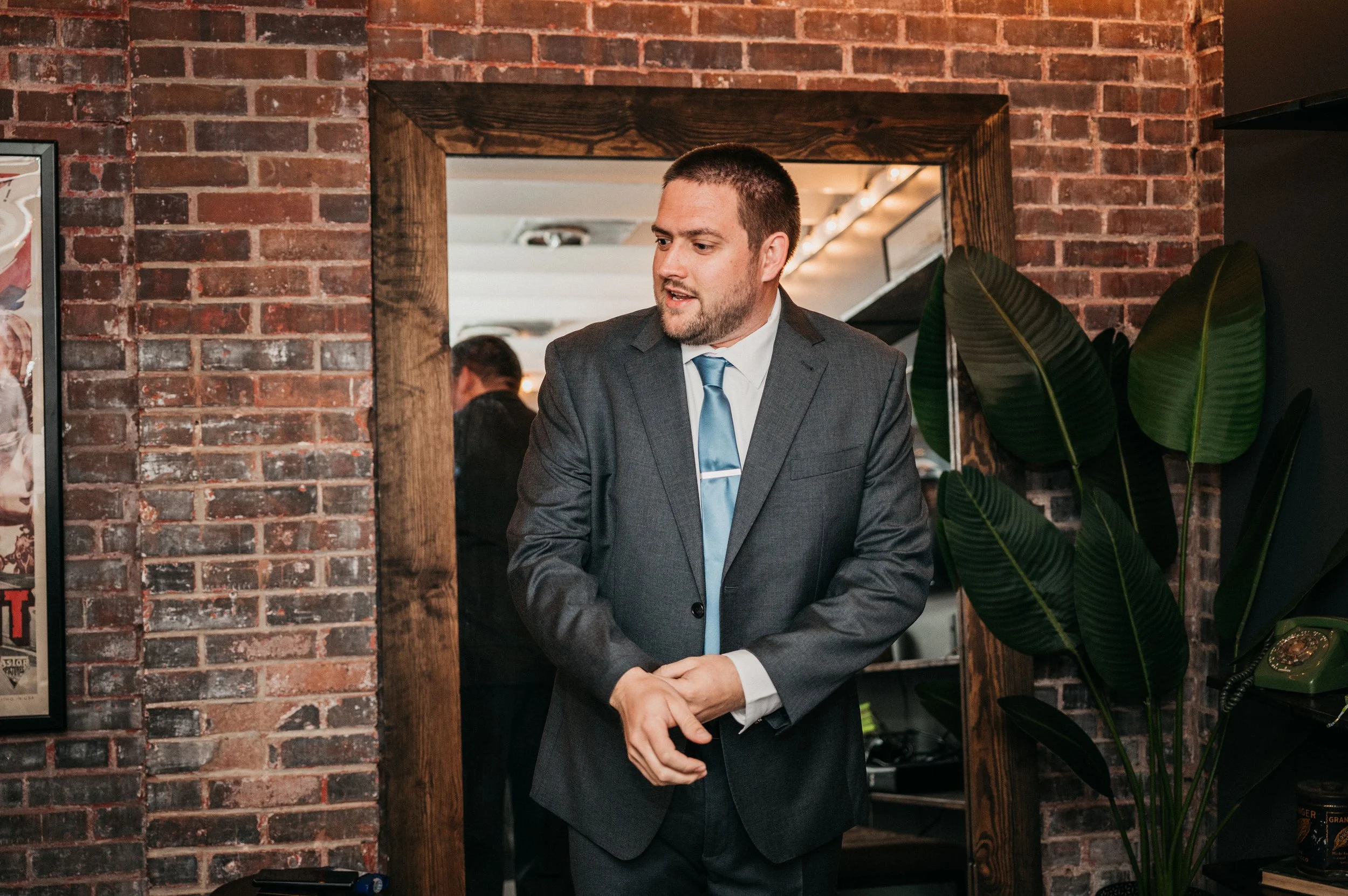 A man in a gray suit and light blue tie standing in front of a mirror with brick wall background, adjusting his sleeve.